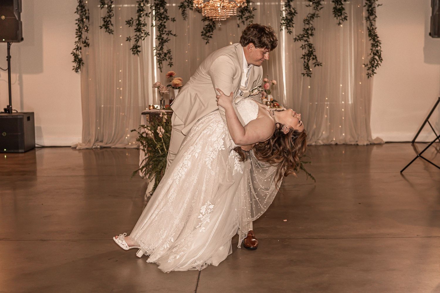 A couple shares a romantic dance dip during their wedding reception on a wooden dance floor with fairy lights and greenery.