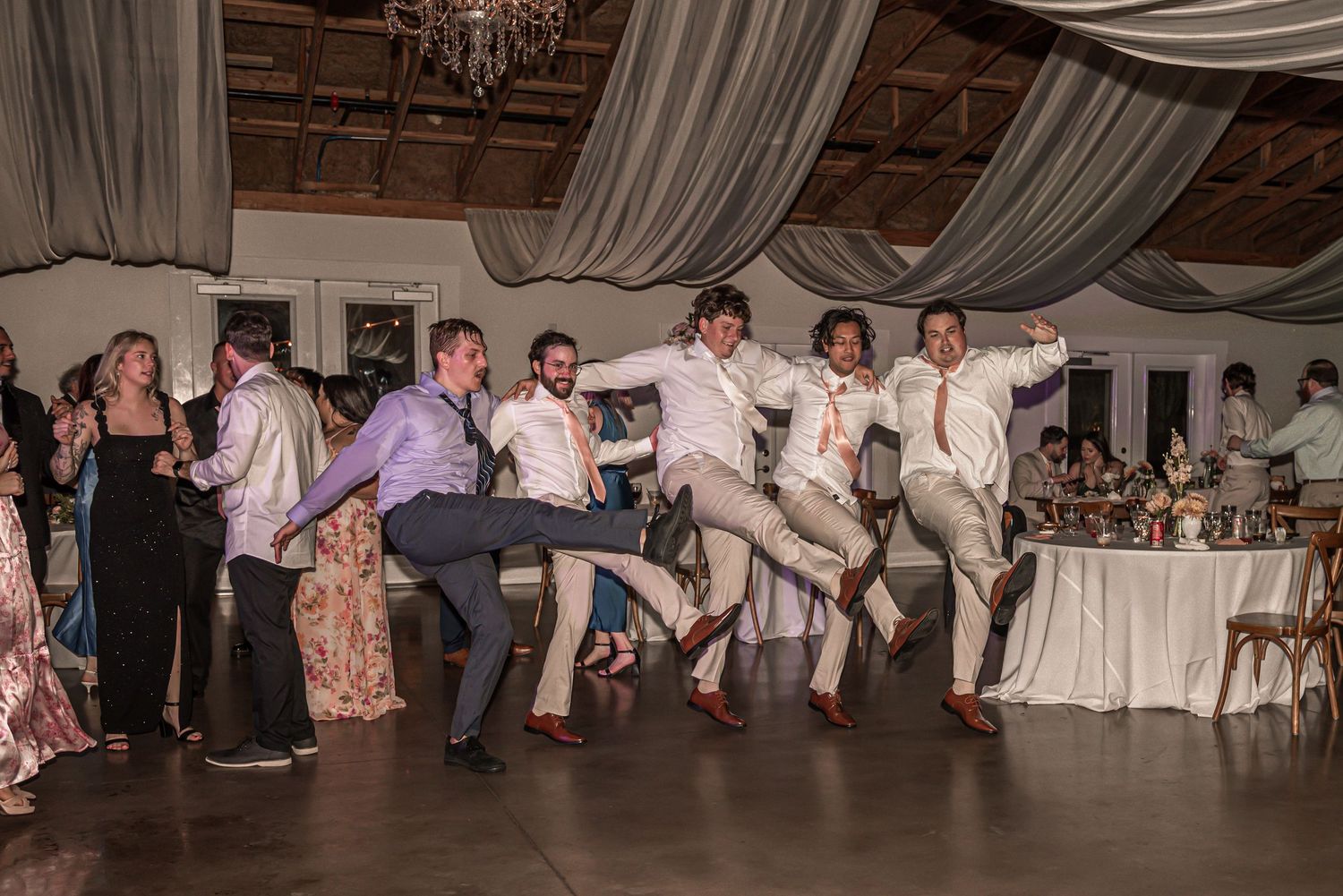 A group of wedding guests performing a synchronized dance move during a reception at an elegant venue with draping decor.