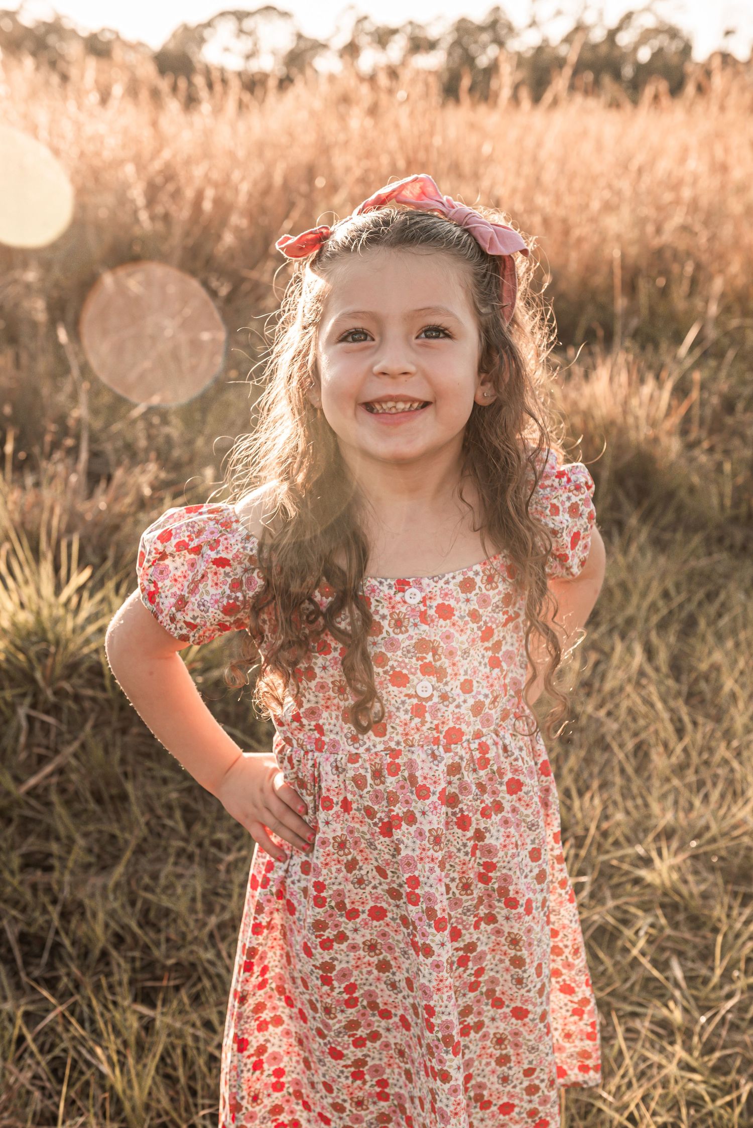 A young child wearing a floral print dress and hair bow poses in a sunlit field at golden hour with glowing bokeh effects.