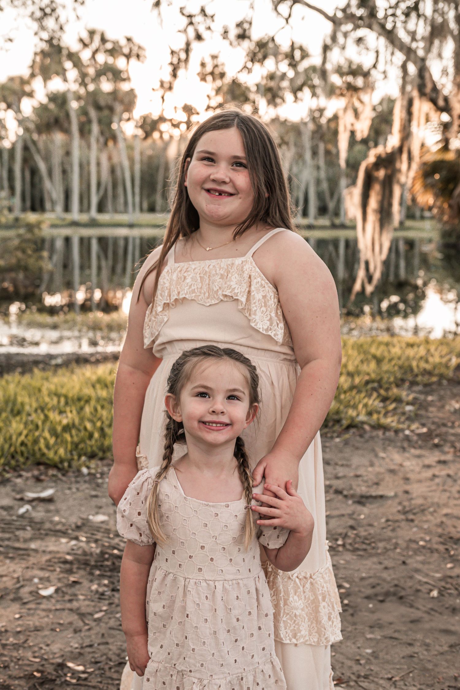 Two children in cream-colored lace dresses pose together outdoors at sunset with trees and a lake in background.