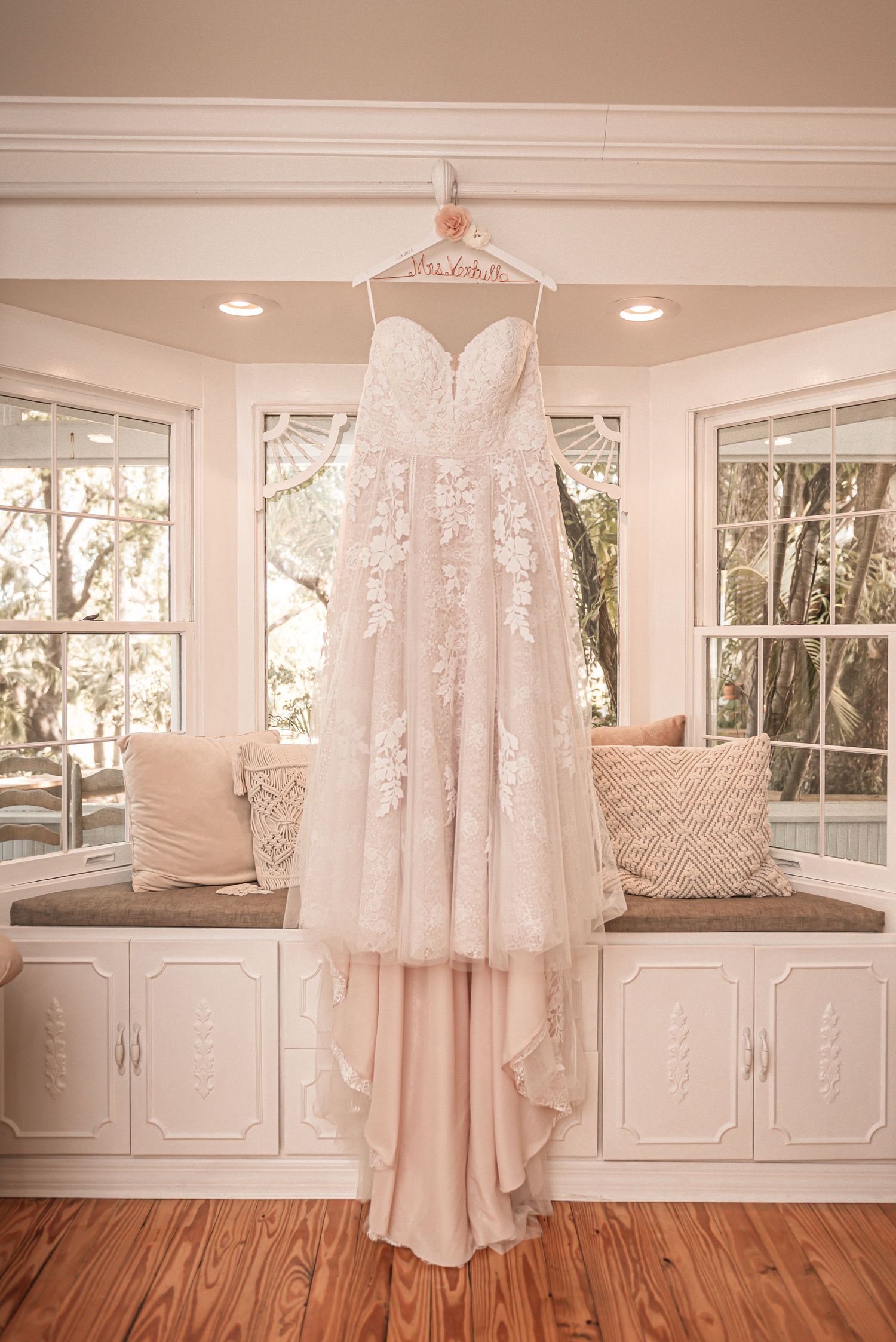 Elegant white lace wedding dress with sweetheart neckline hanging in sunlit bay window against cream walls.
