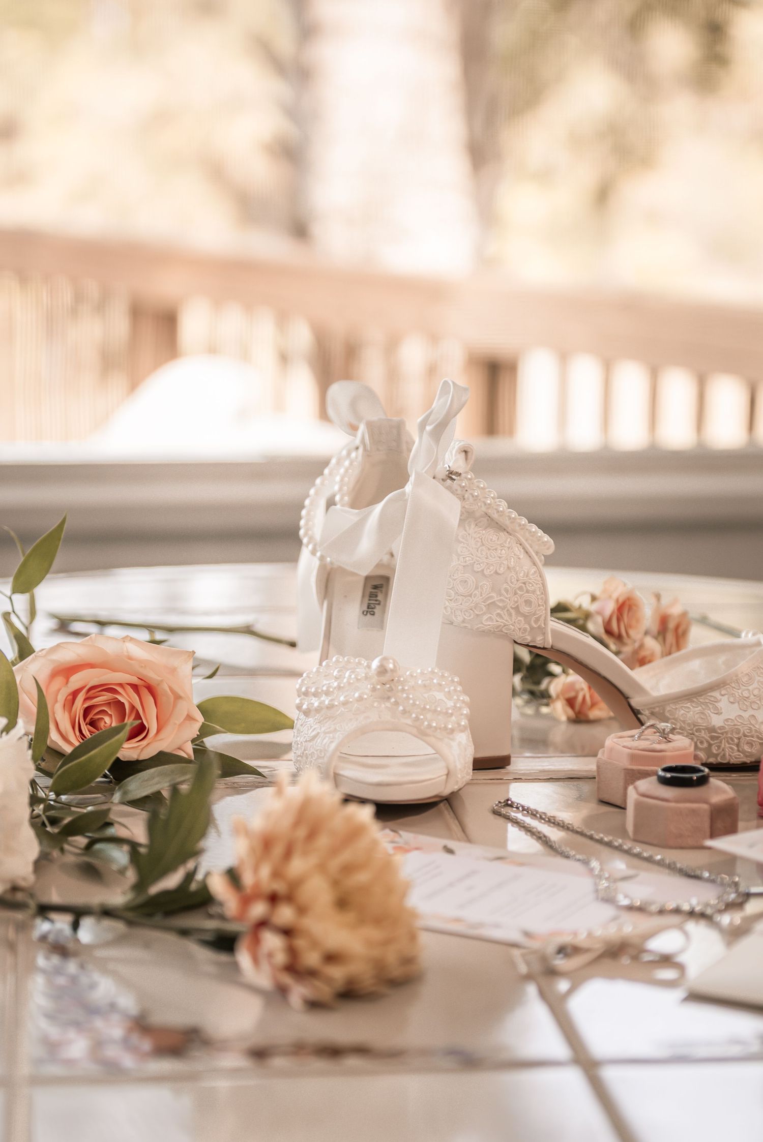 Elegant white lace bridal shoes displayed with pink roses and wedding accessories on a light table.