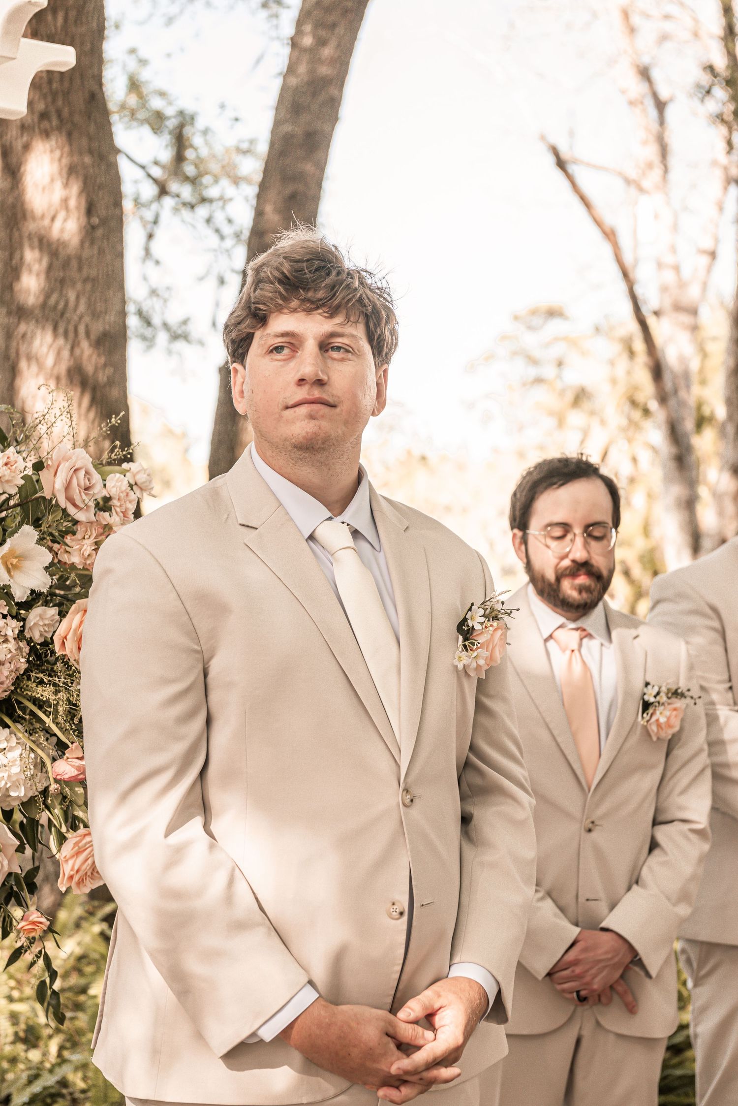 Two groomsmen in beige suits with peach accents stand outdoors among trees at a wedding ceremony.