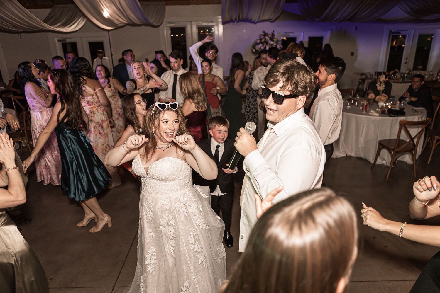 Wedding guests celebrate and dance on a crowded dance floor at an indoor reception with purple lighting.