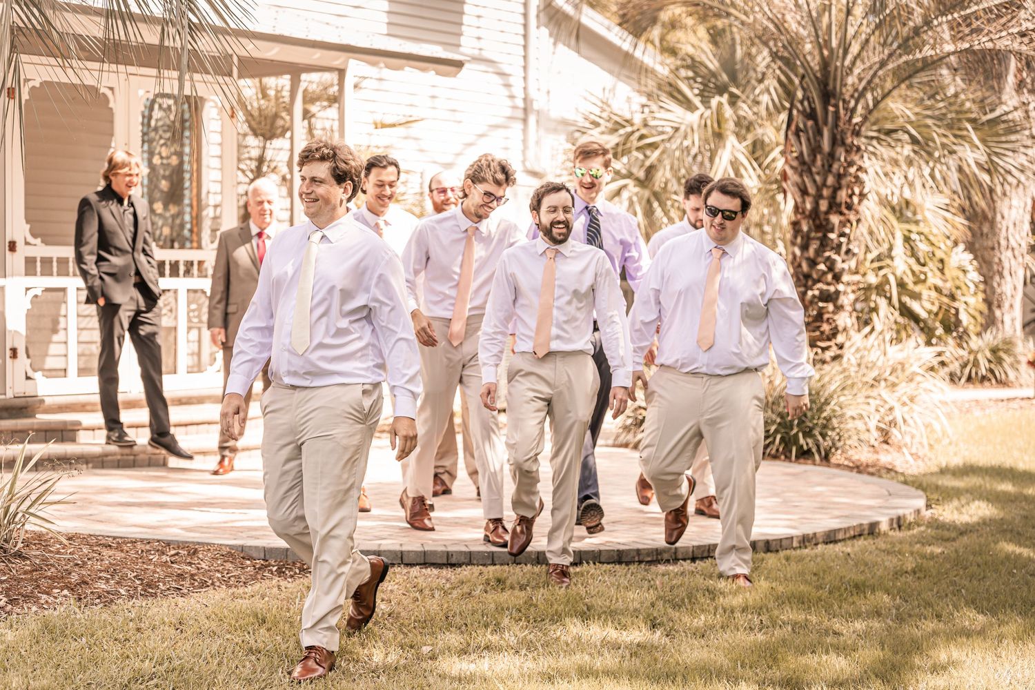 A group of groomsmen in light colored attire walk together outdoors on a sunny day near a palm tree.