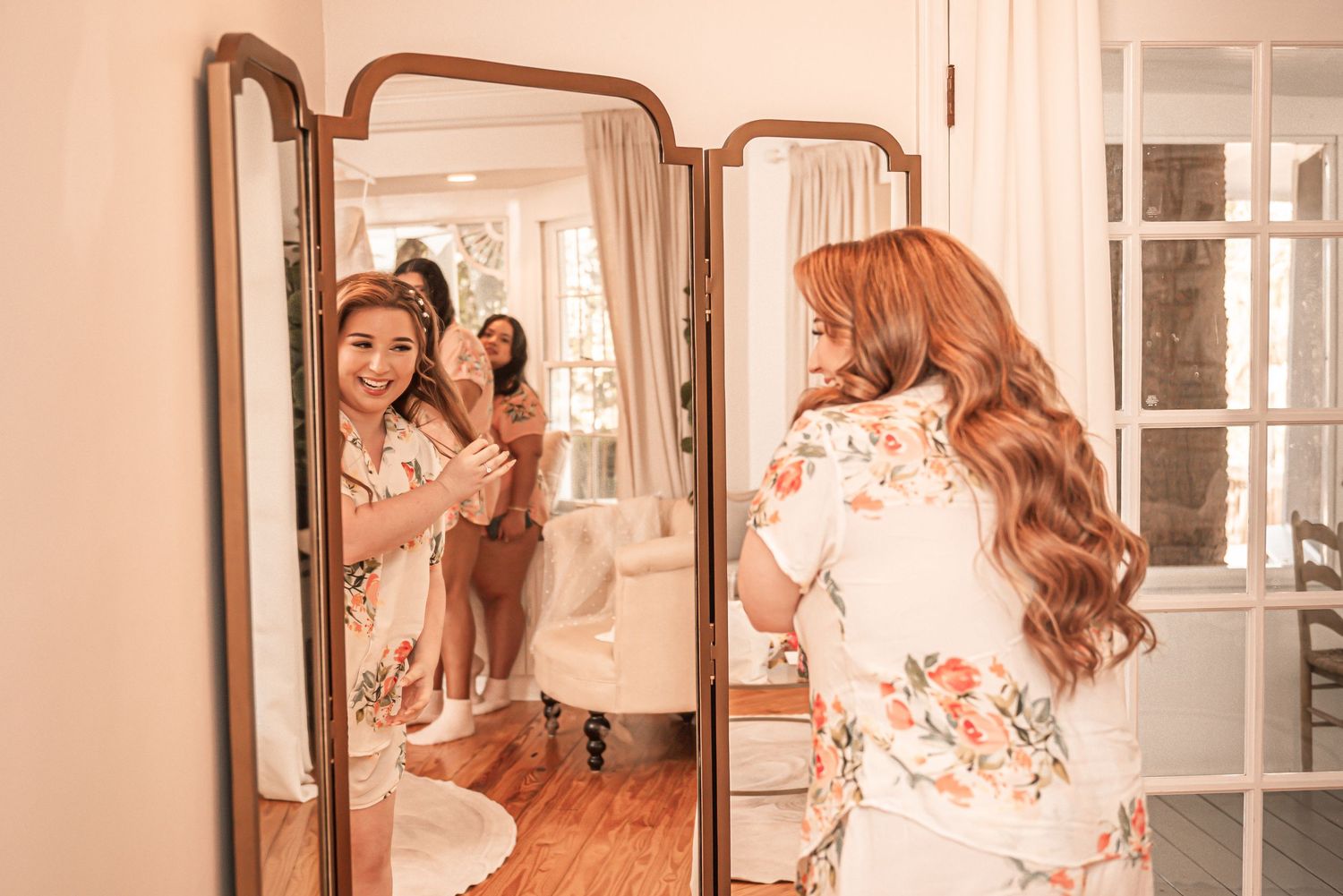 A figure in a floral robe stands in front of an elegant tri-fold mirror in a warm peachy-toned room.