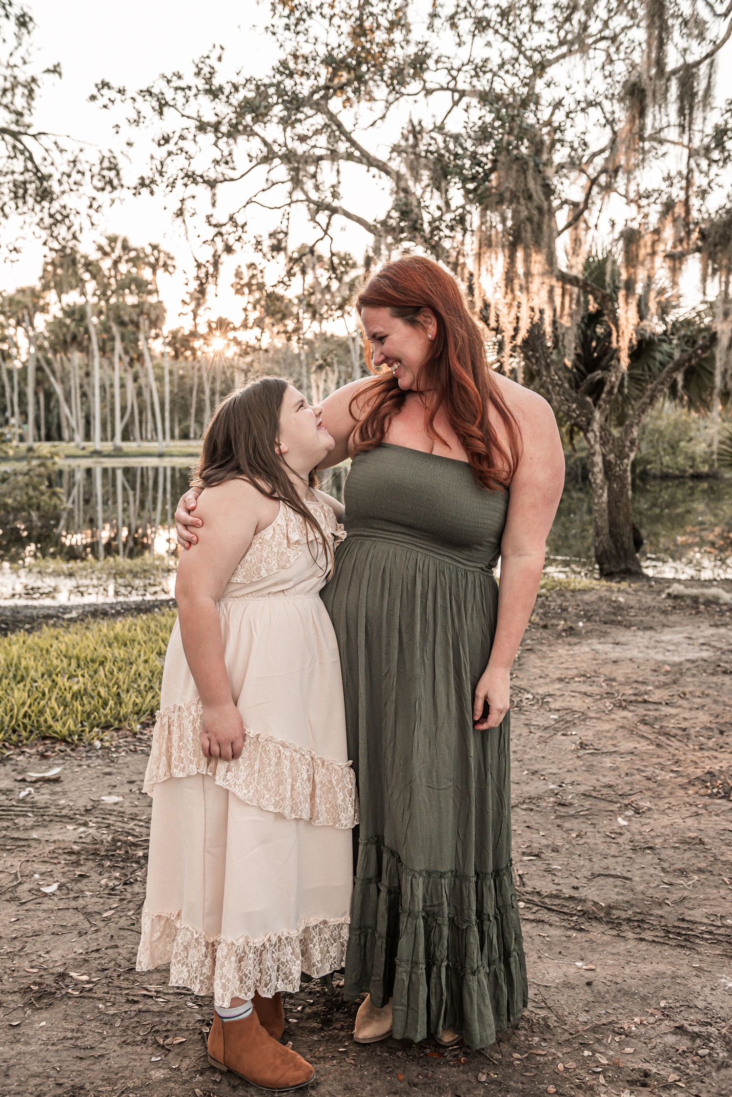 Two people in maxi dresses standing together at sunset near a lake with Spanish moss draped trees in background.