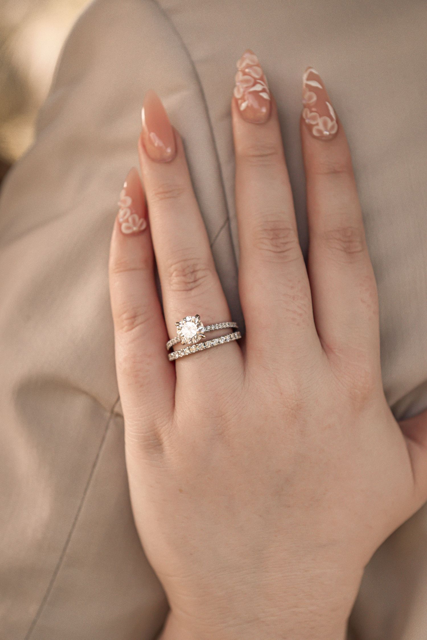 Hand wearing an elegant engagement ring and wedding band set with floral nail art designs on pale pink manicured nails.