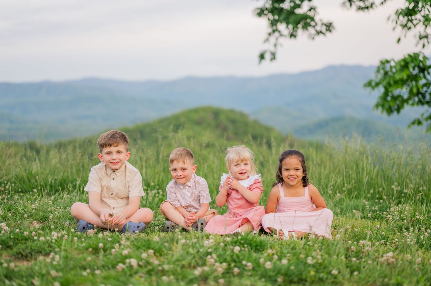 Children sit together in a line on a grassy hilltop with mountains and trees visible in the background.