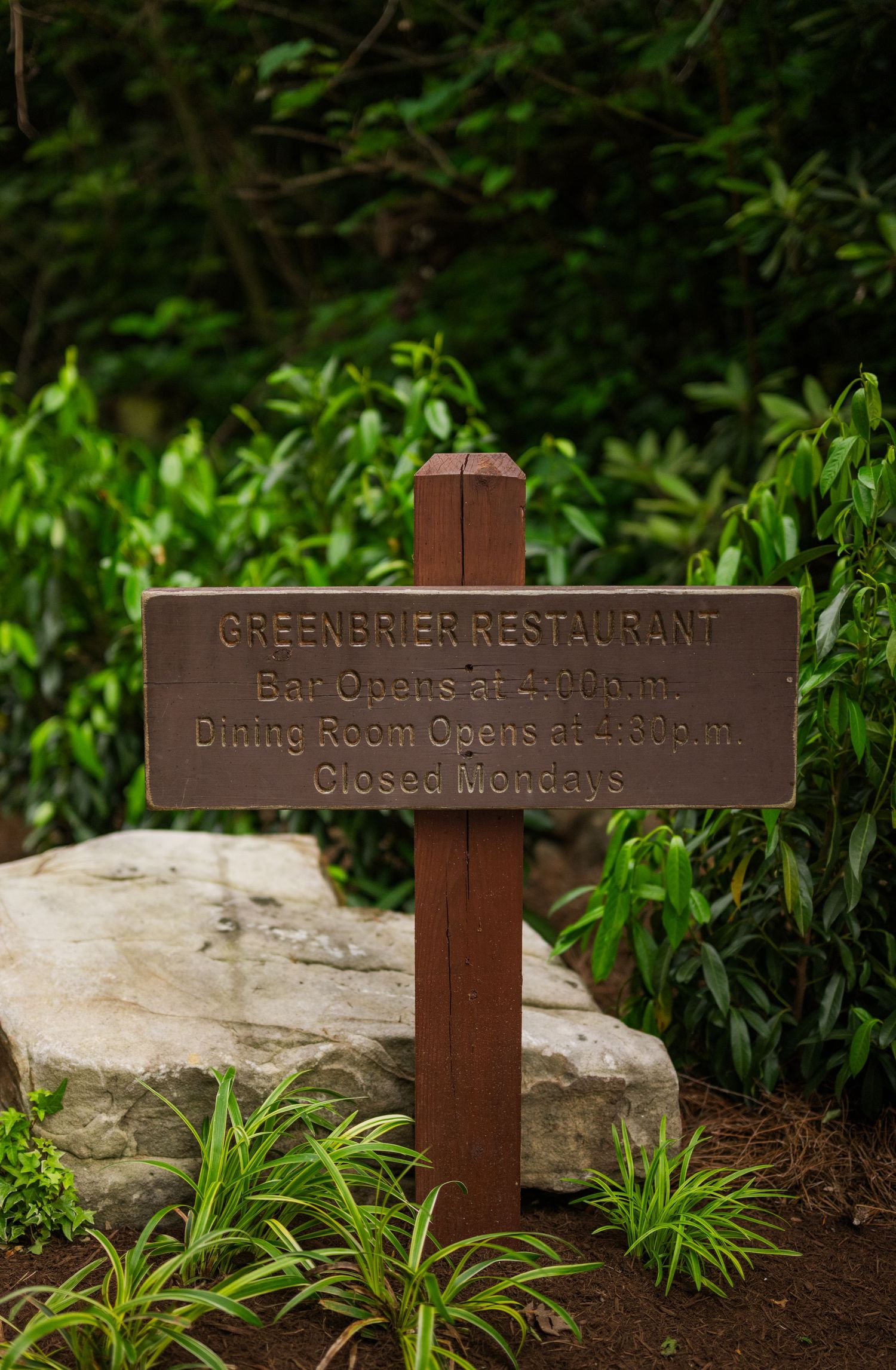 A wooden sign for Greenbrier Restaurant mounted on a post in a garden setting with green foliage and rocks.
