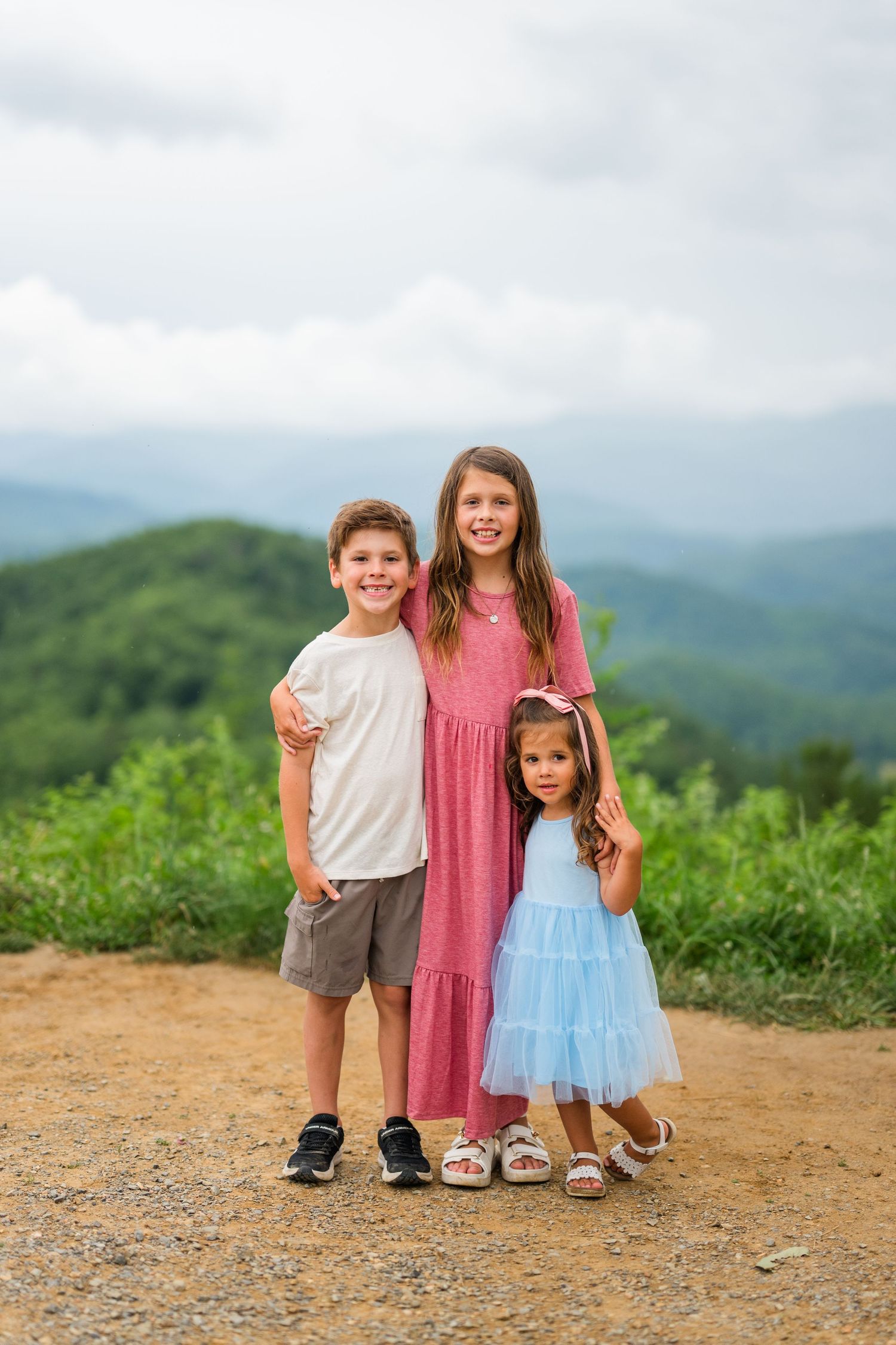 Three children stand on a mountain trail with green hills and misty mountains in the scenic background.