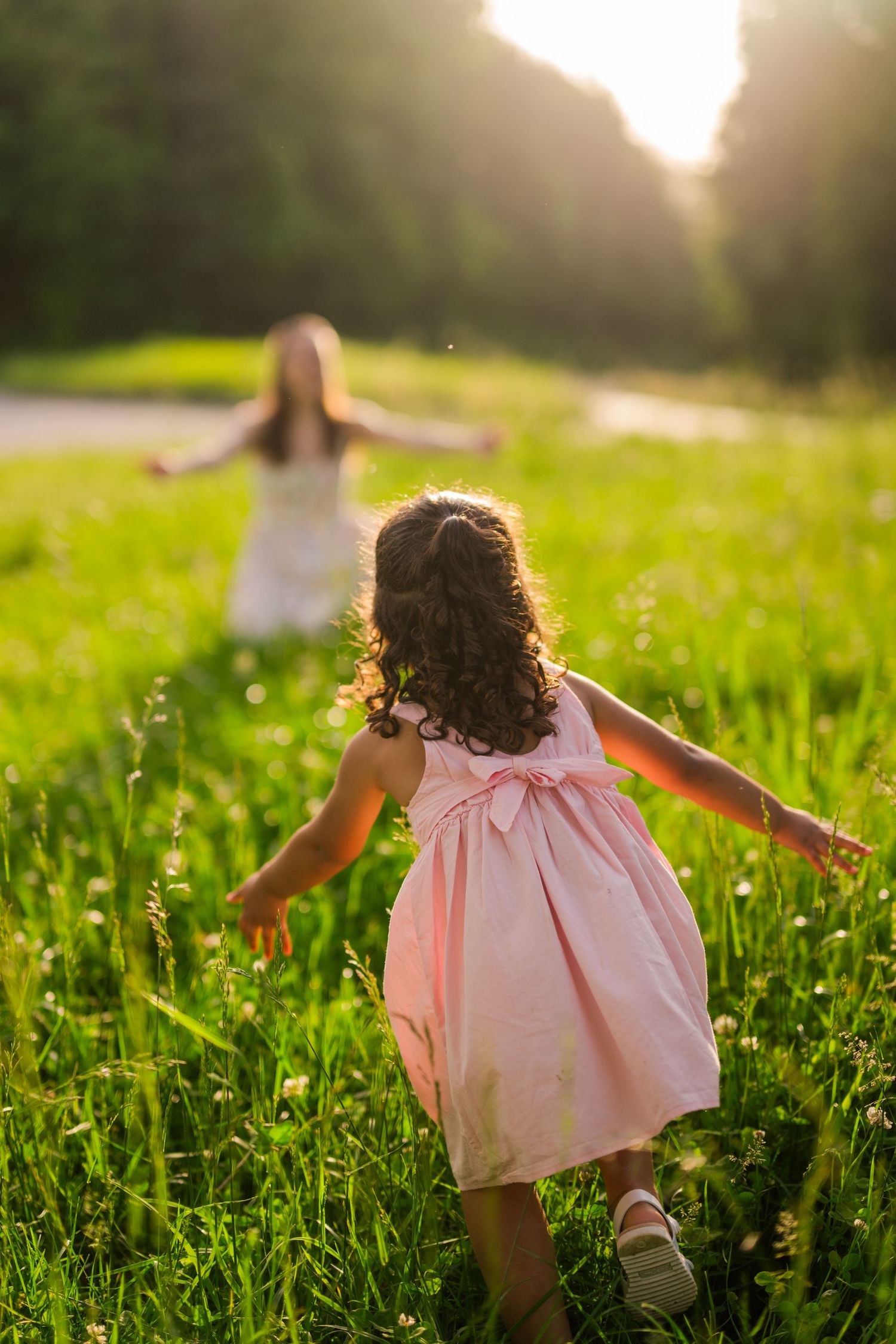 Two figures in dresses running through a sunlit meadow with tall grass at golden hour.