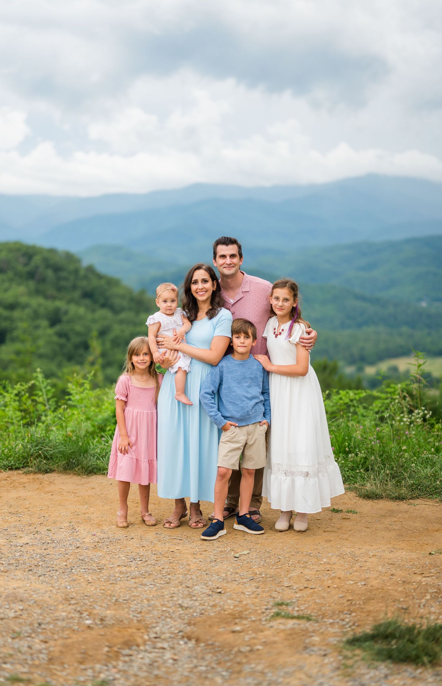 A scenic family portrait captured on a mountaintop trail with rolling blue mountains and cloudy skies in the background.
