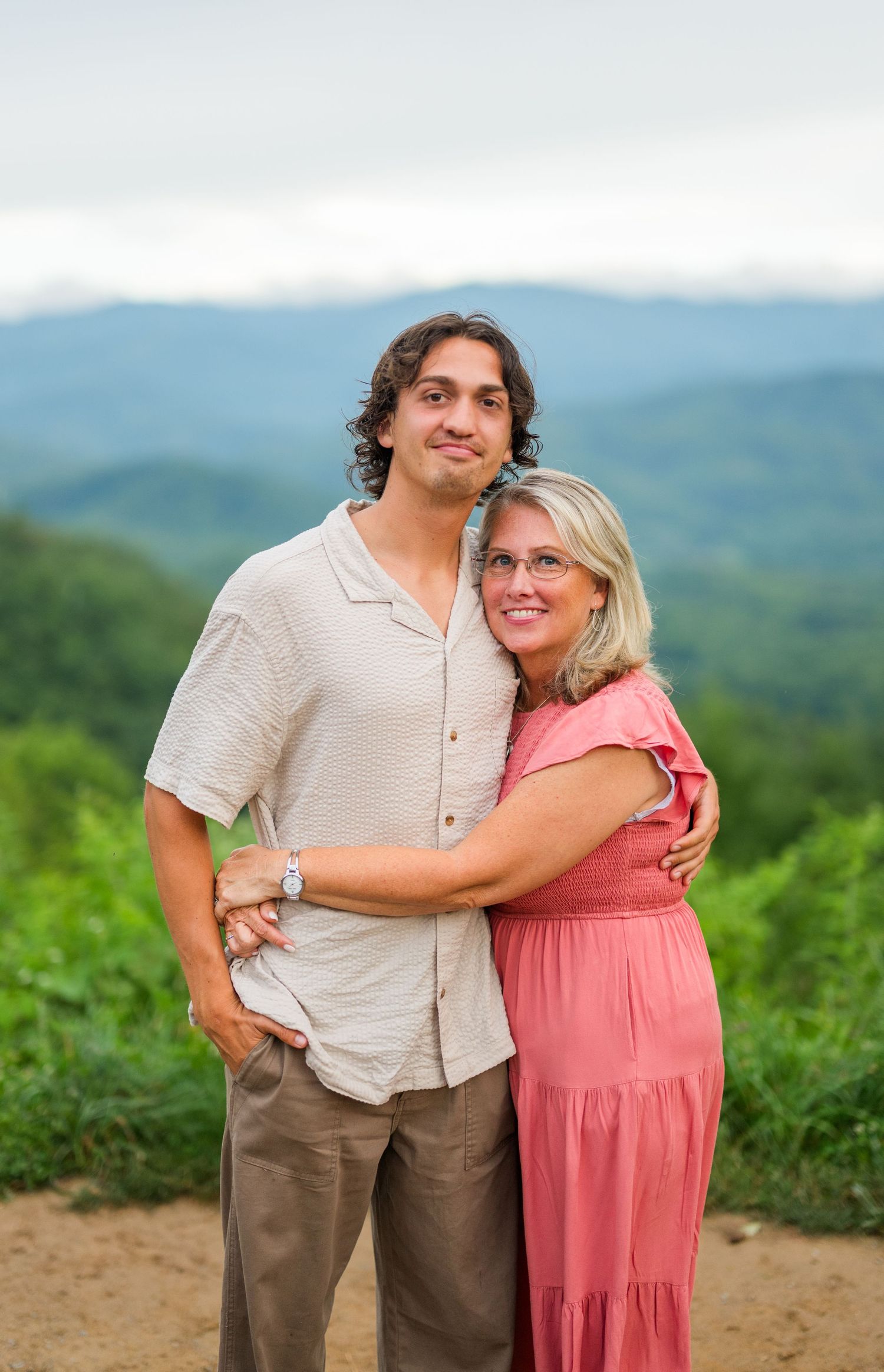 A couple in casual attire embraces while standing on a mountain overlook with green hills and blue skies in the background.