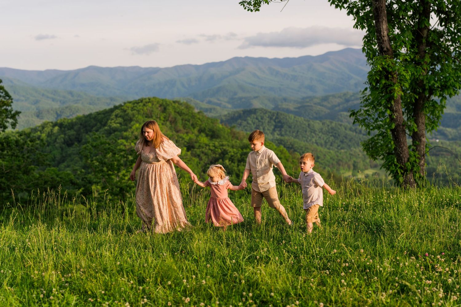 A group holds hands while running through a lush mountain meadow at sunset, wearing flowing dresses against rolling hills.
