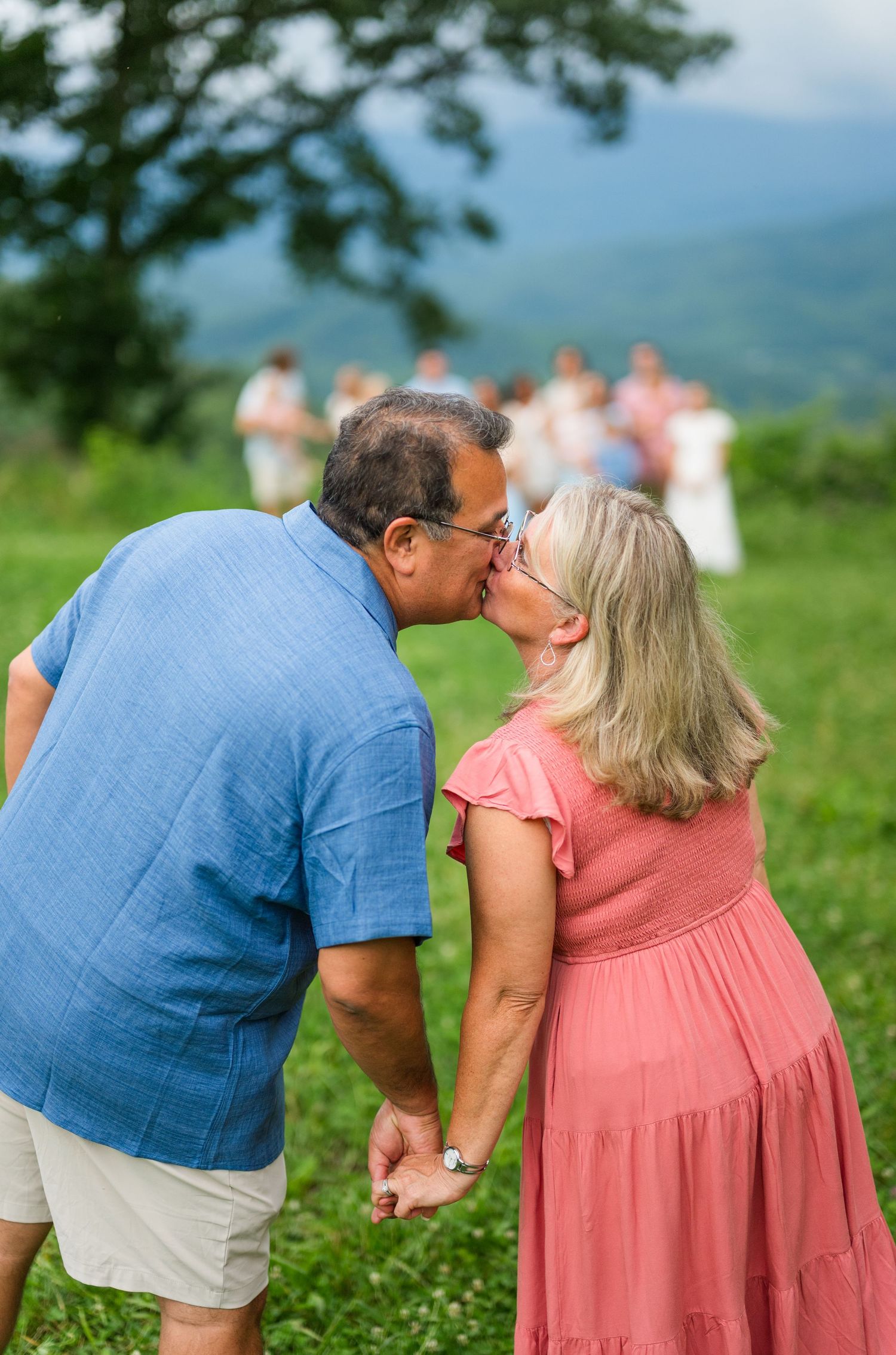 A couple in blue and pink attire share a tender moment on a grassy hilltop with mountains in the background.