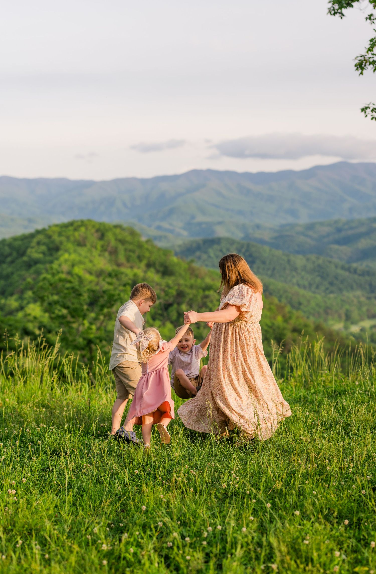A joyful family dances together on a hilltop meadow with rolling mountains in the background at sunset.
