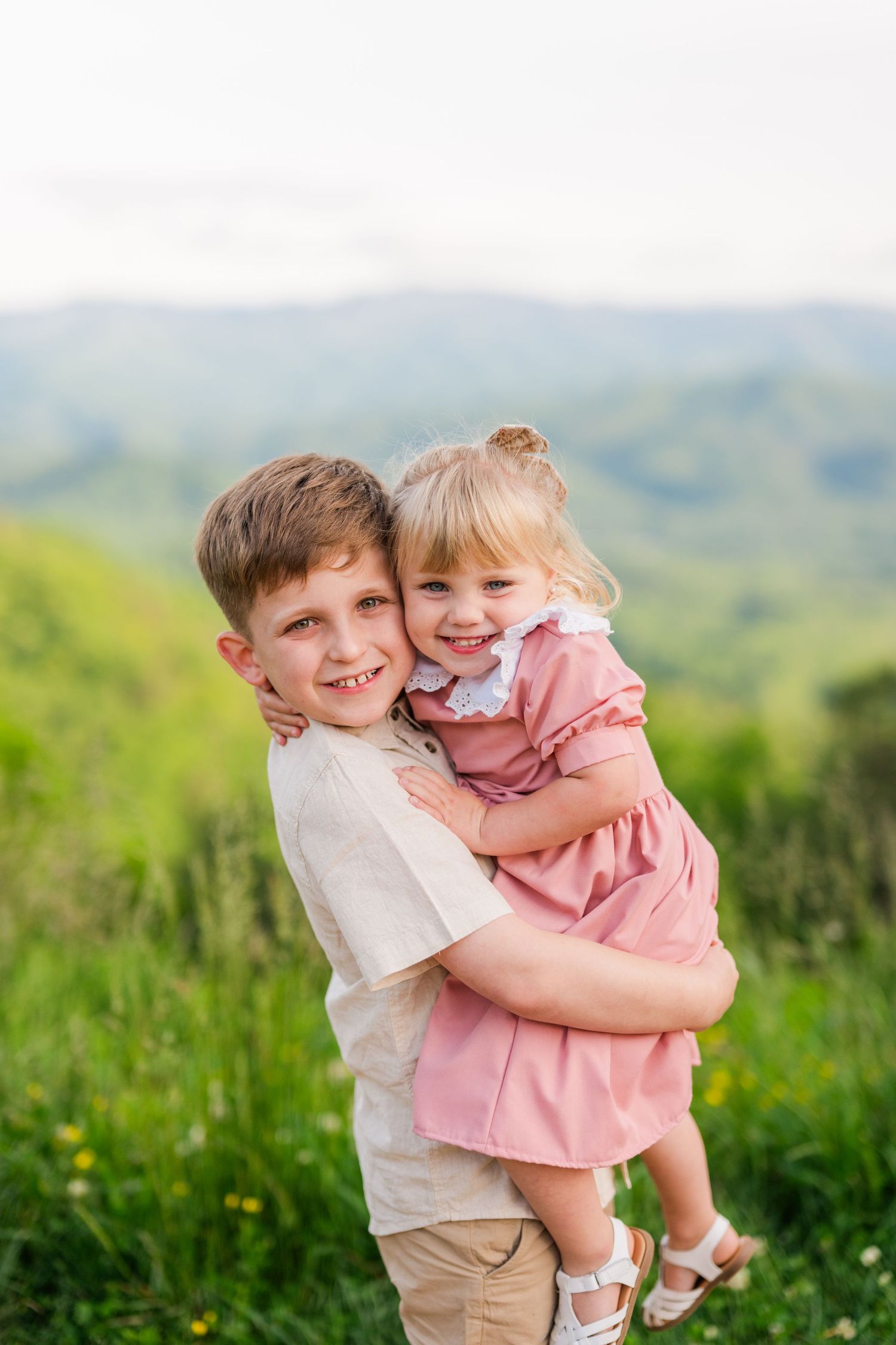 Two children share a joyful embrace while standing in a grassy meadow with rolling hills in the background off of the Foothills Parkway