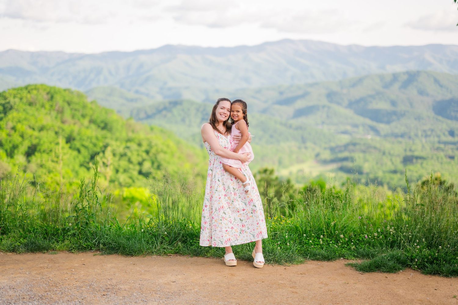 Two people embrace on a scenic mountain trail overlooking green rolling hills and distant mountains on a sunny day.