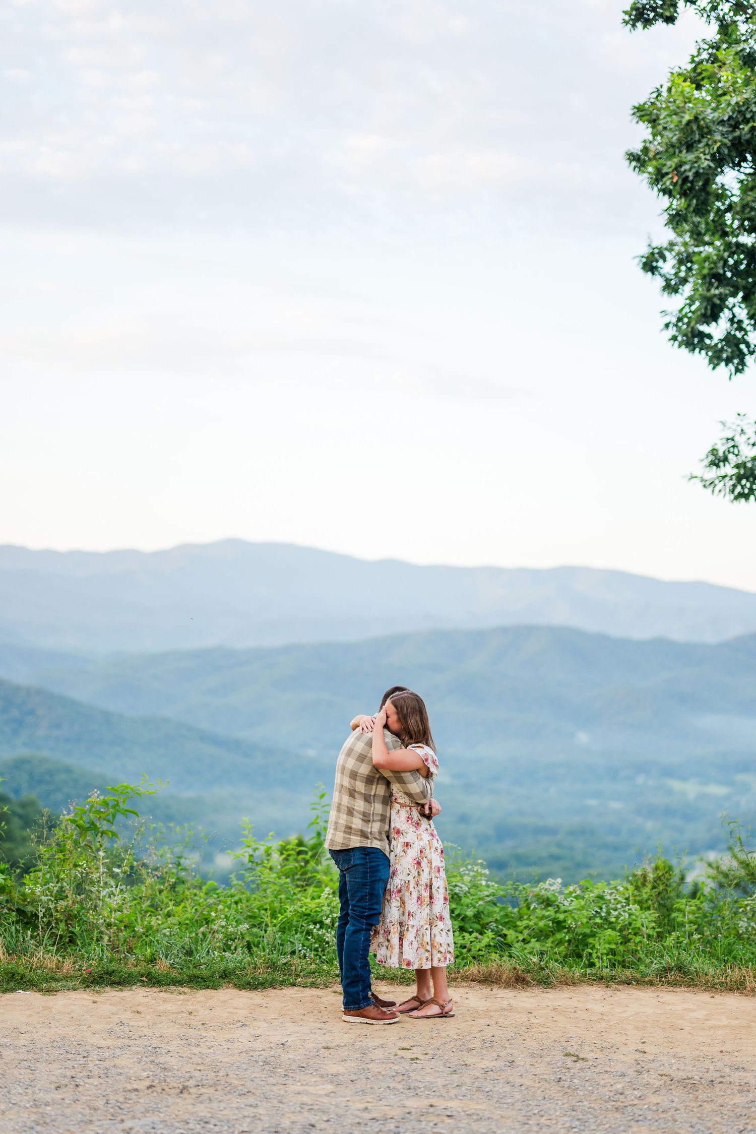 A romantic couple shares a tender moment on a hilltop overlook with scenic mountains and forests in the background.