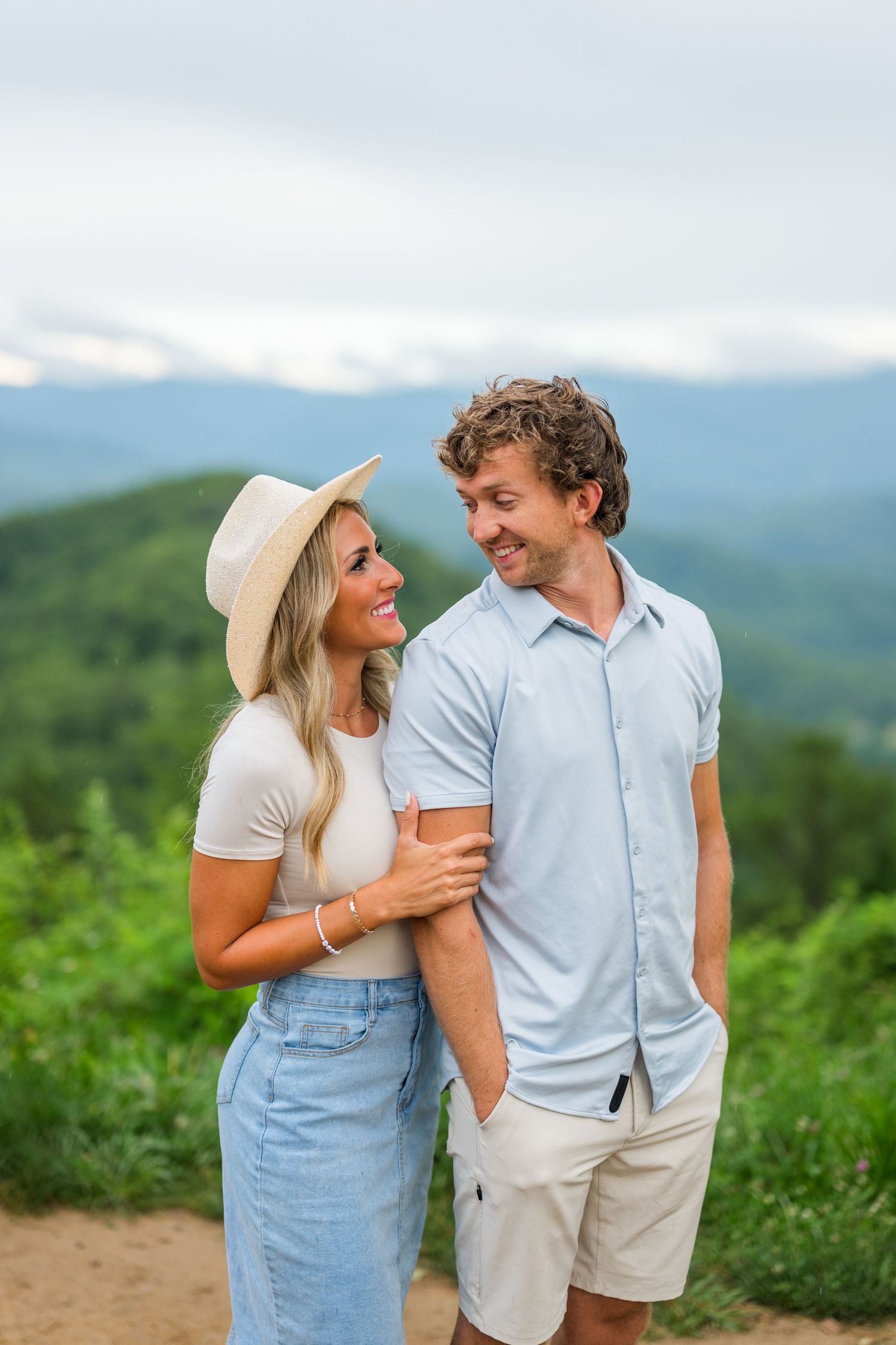 A couple shares a warm moment together while hiking on a mountain trail with scenic views in the background.