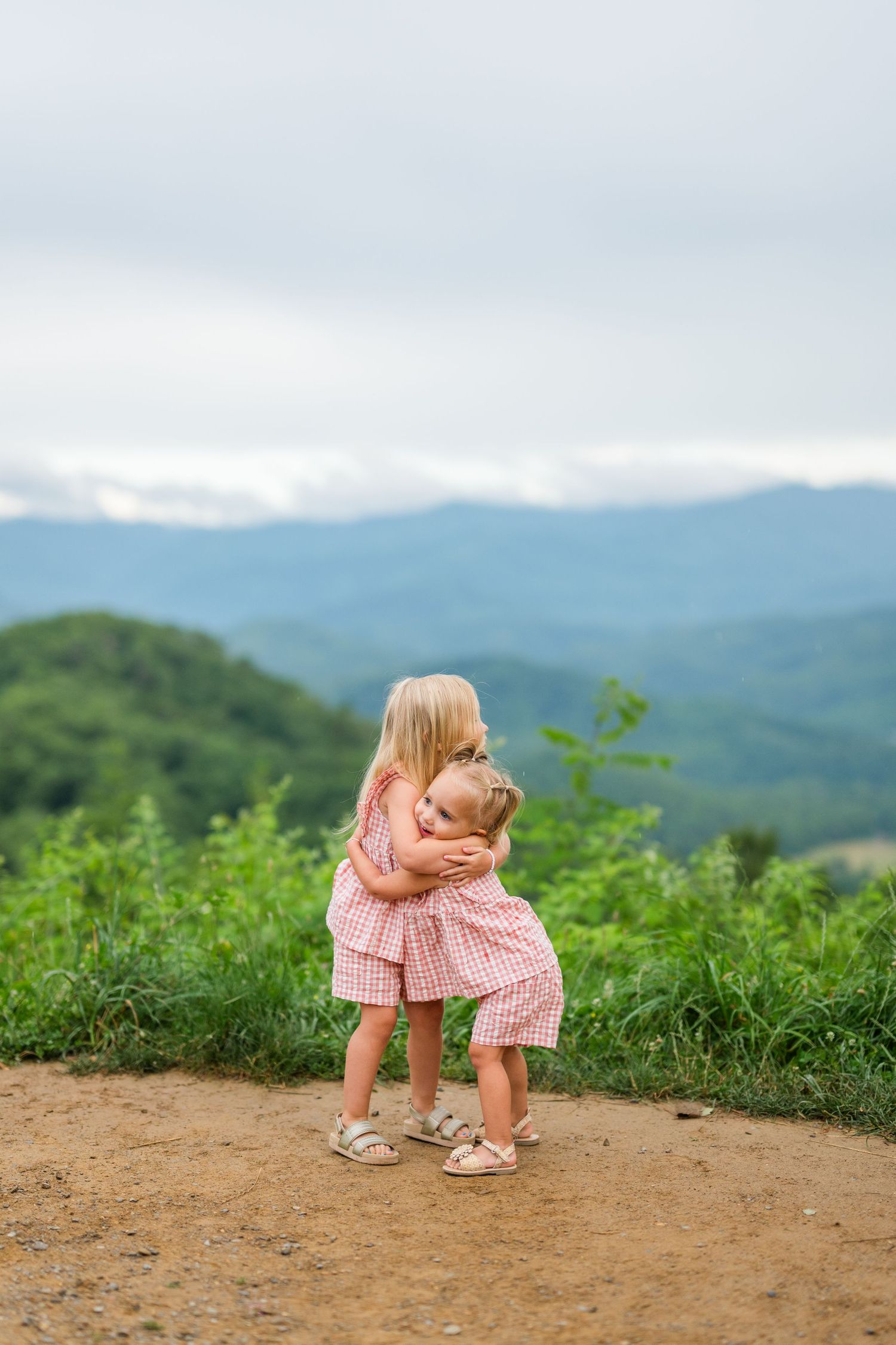 Small child in pink gingham dress hugs teddy bear while standing on mountaintop trail with misty peaks in background.