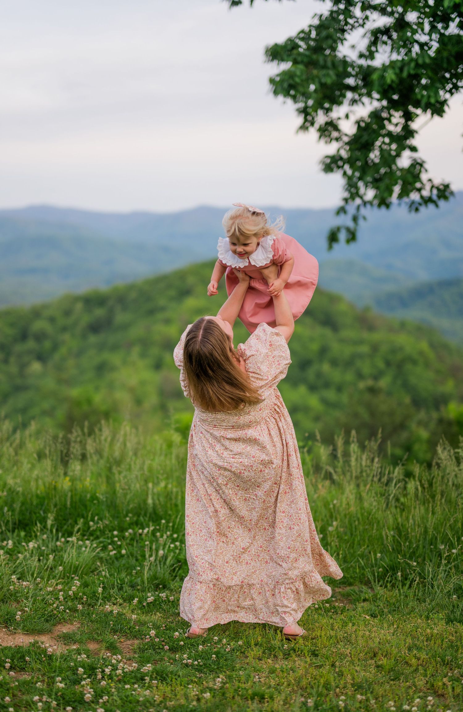 Two figures in floral dresses share a tender moment on a hilltop overlooking a mountain landscape at sunset.