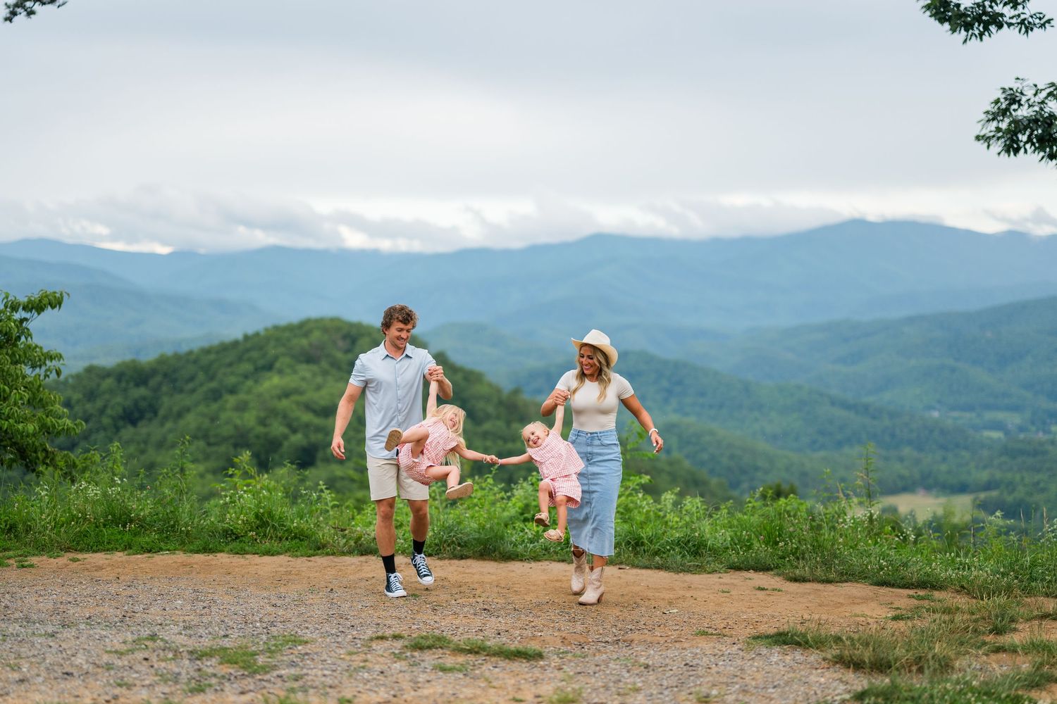 Two people hold hands while running along a mountain trail with scenic Blue Ridge mountains in the background.