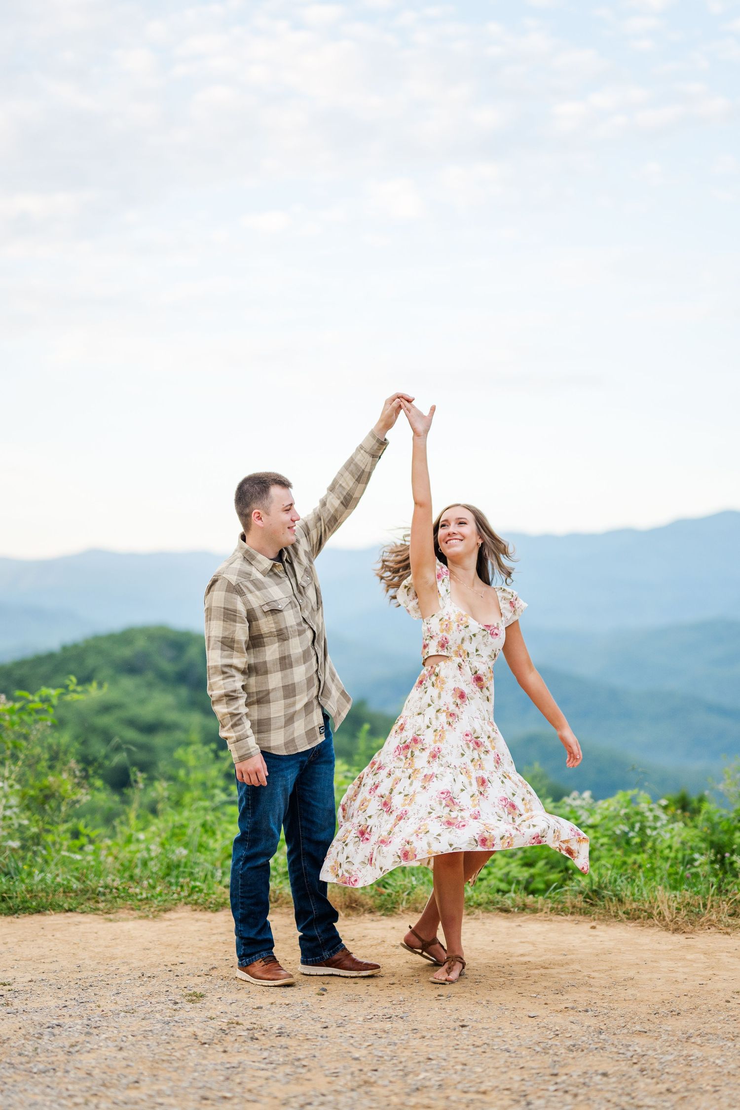 Couple dancing joyfully on a mountaintop trail with green mountains and misty valleys in the background.