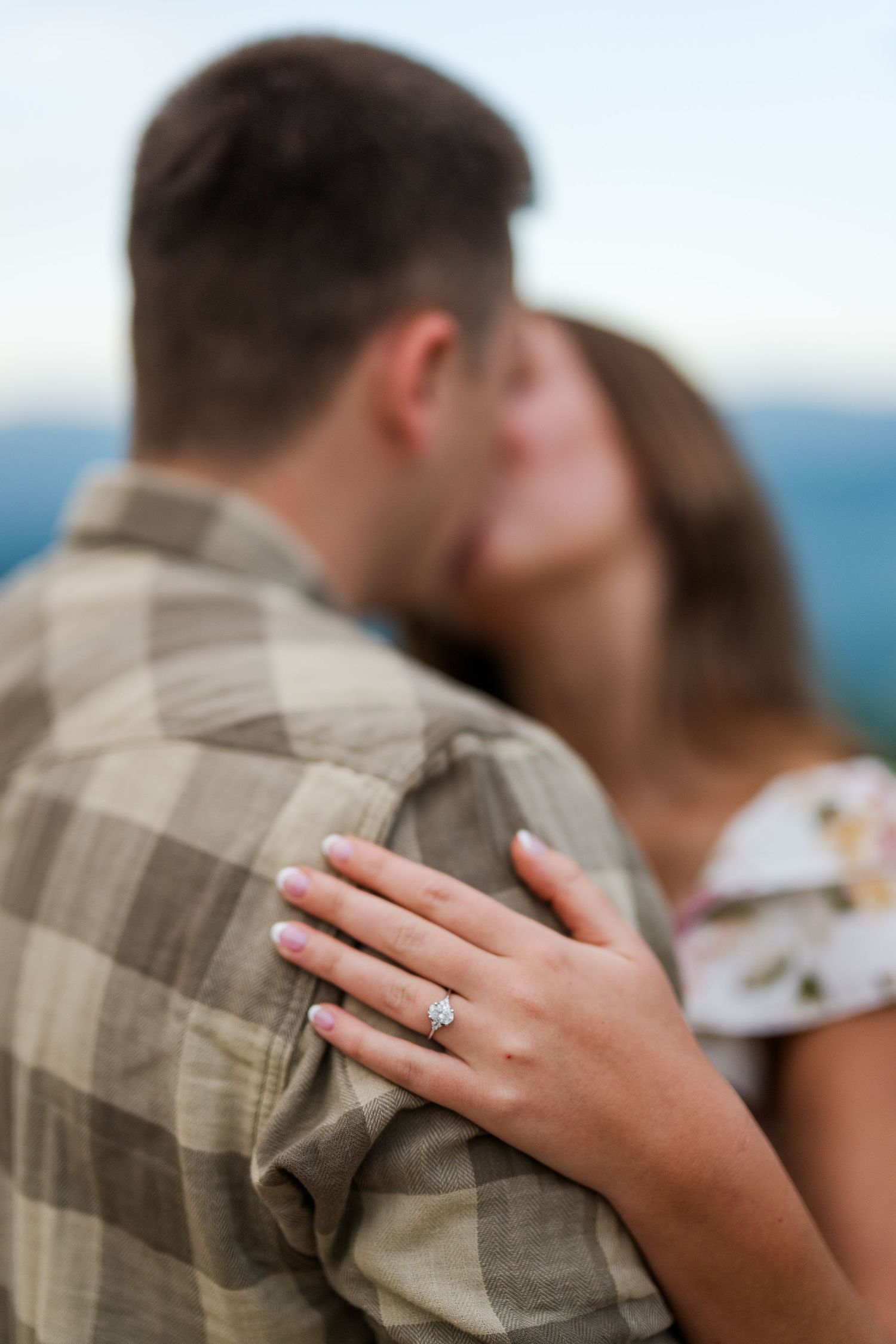 A close-up shot of an engagement ring on a woman's hand resting on someone wearing a plaid shirt outdoors.