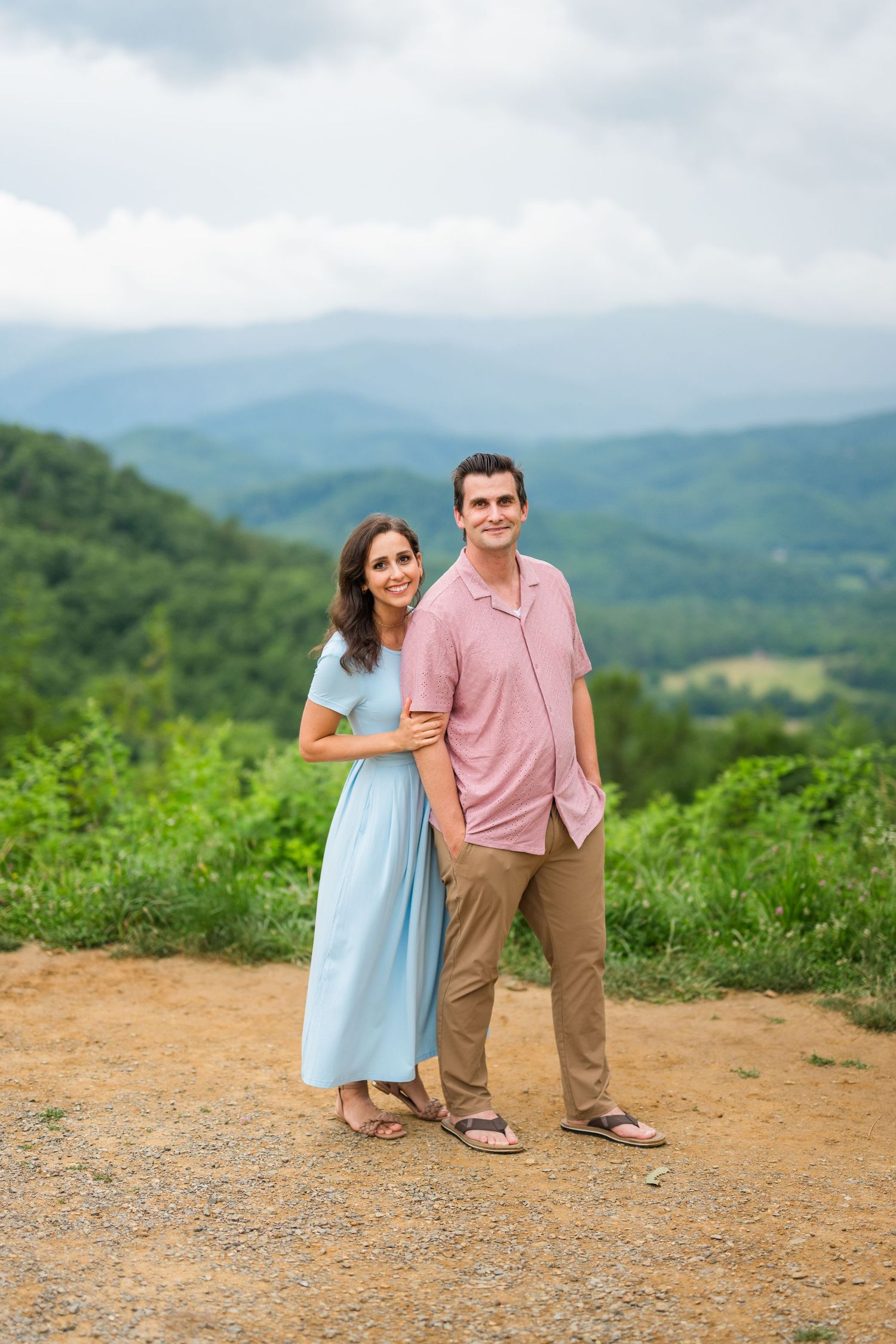 A couple stands together on a mountain overlook with a scenic vista of green rolling hills and mountains in the background.