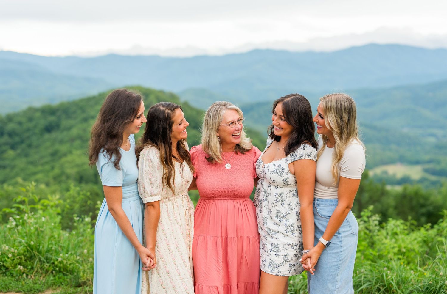 A group of friends in summer dresses share joyful laughter together on a scenic mountaintop overlook with rolling hills behind them.