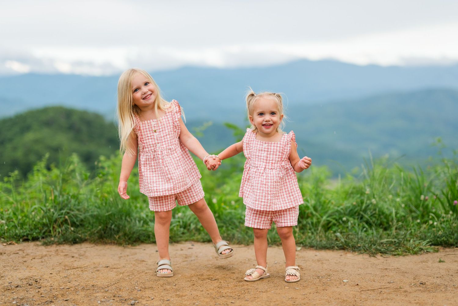 Two people in matching pink outfits walk along a mountain path with scenic mountain views in the background.