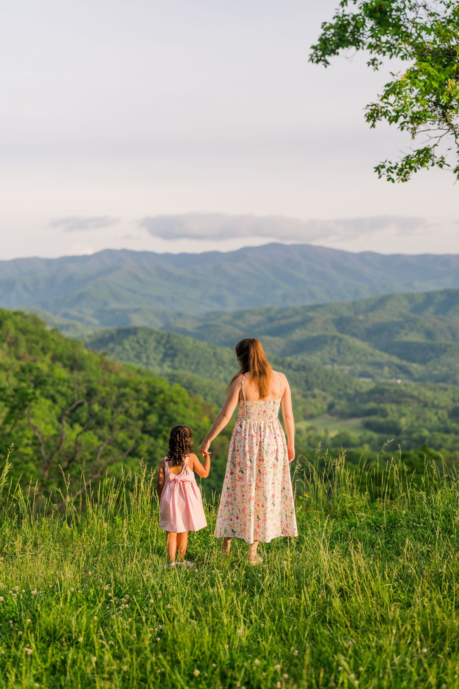 Two figures in floral dresses stand holding hands while overlooking a scenic mountain landscape on a grassy hilltop.