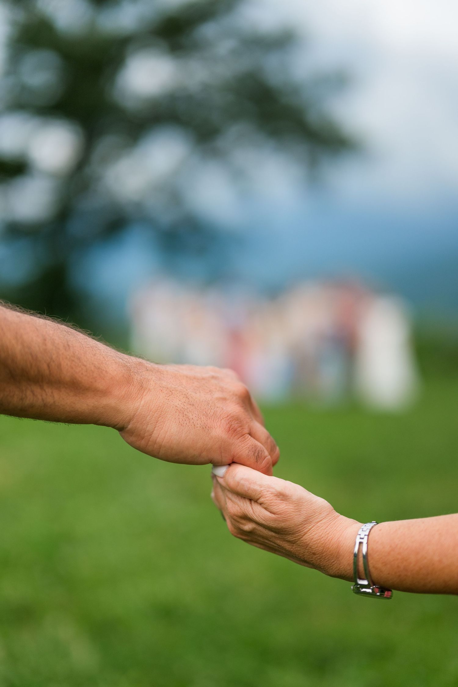 Two hands reach out to hold each other against a blurred outdoor background with grass and sky.