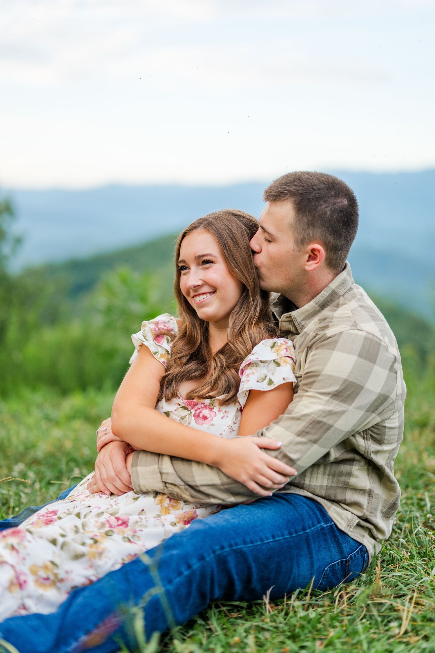 A couple sits together on a grassy hillside overlooking mountain views while sharing an intimate embrace.