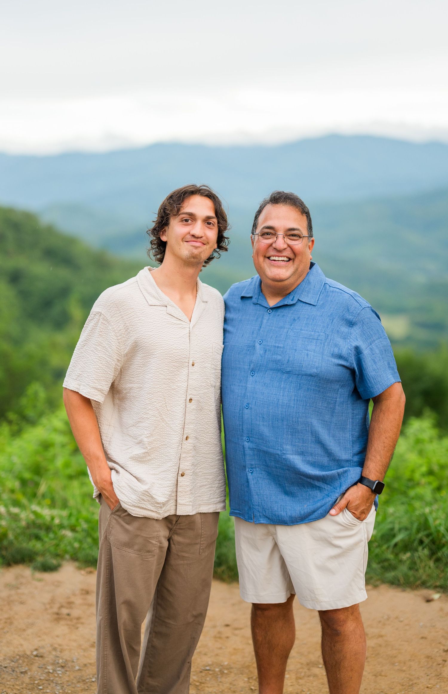 Two people wearing casual attire stand together on a hilltop trail with green mountains in the background.
