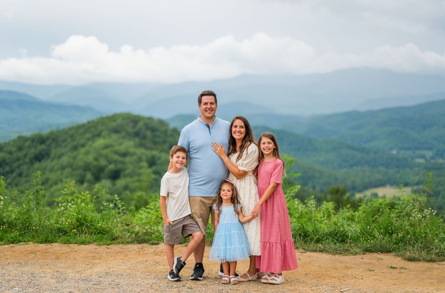 A family enjoys a scenic mountain overlook on a sunny day surrounded by lush green rolling hills and distant peaks.