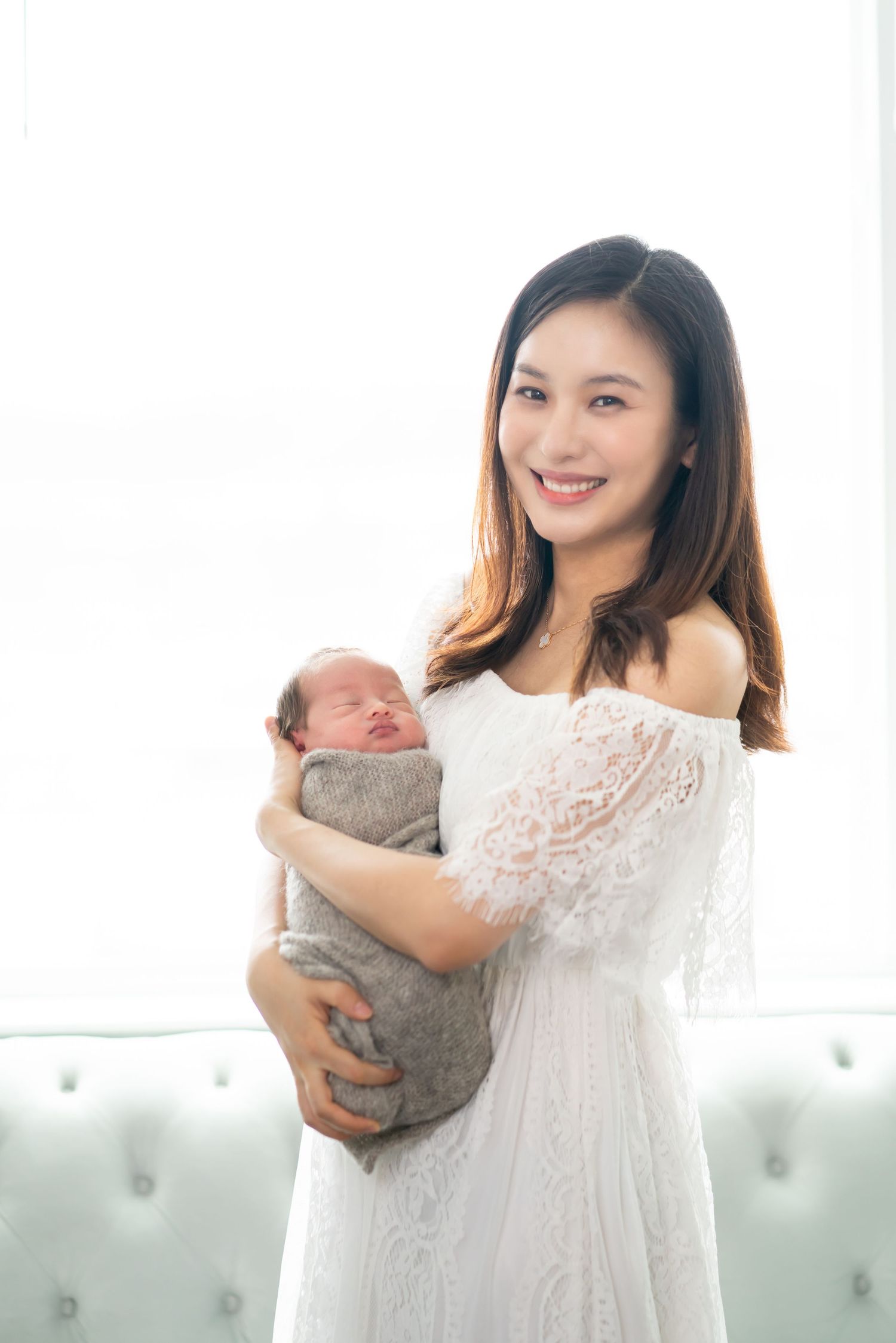 Mother in white lace dress holding newborn baby in bright room.