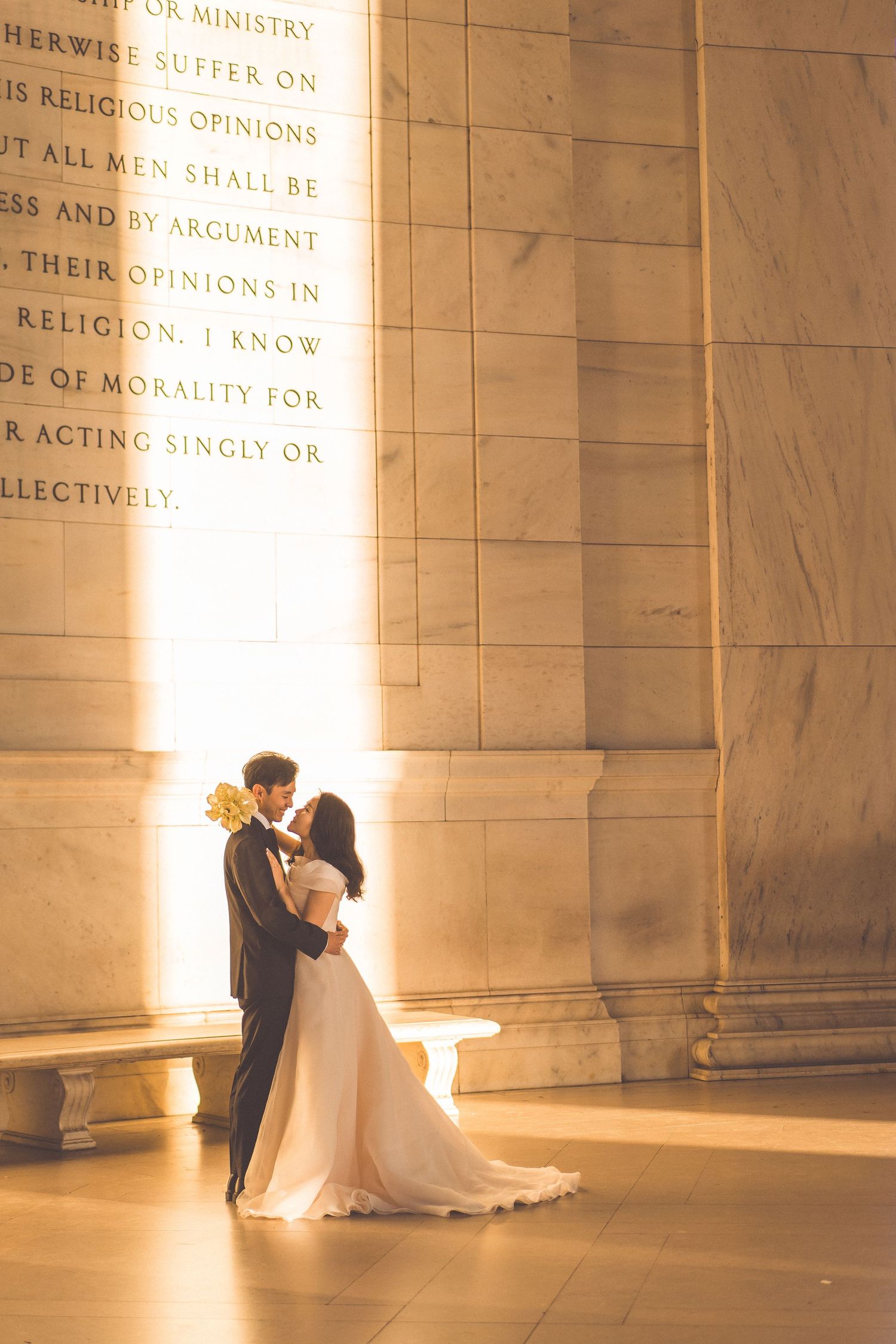Wedding couple silhouetted against illuminated stone wall with carved text at a historic memorial building.