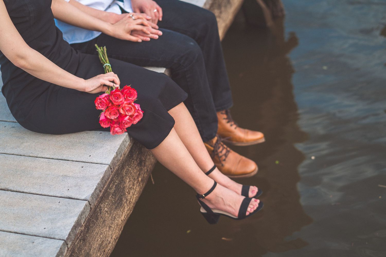 Two people sitting on dock with legs dangling over water, one holding red roses, wearing casual shoes and sandals.