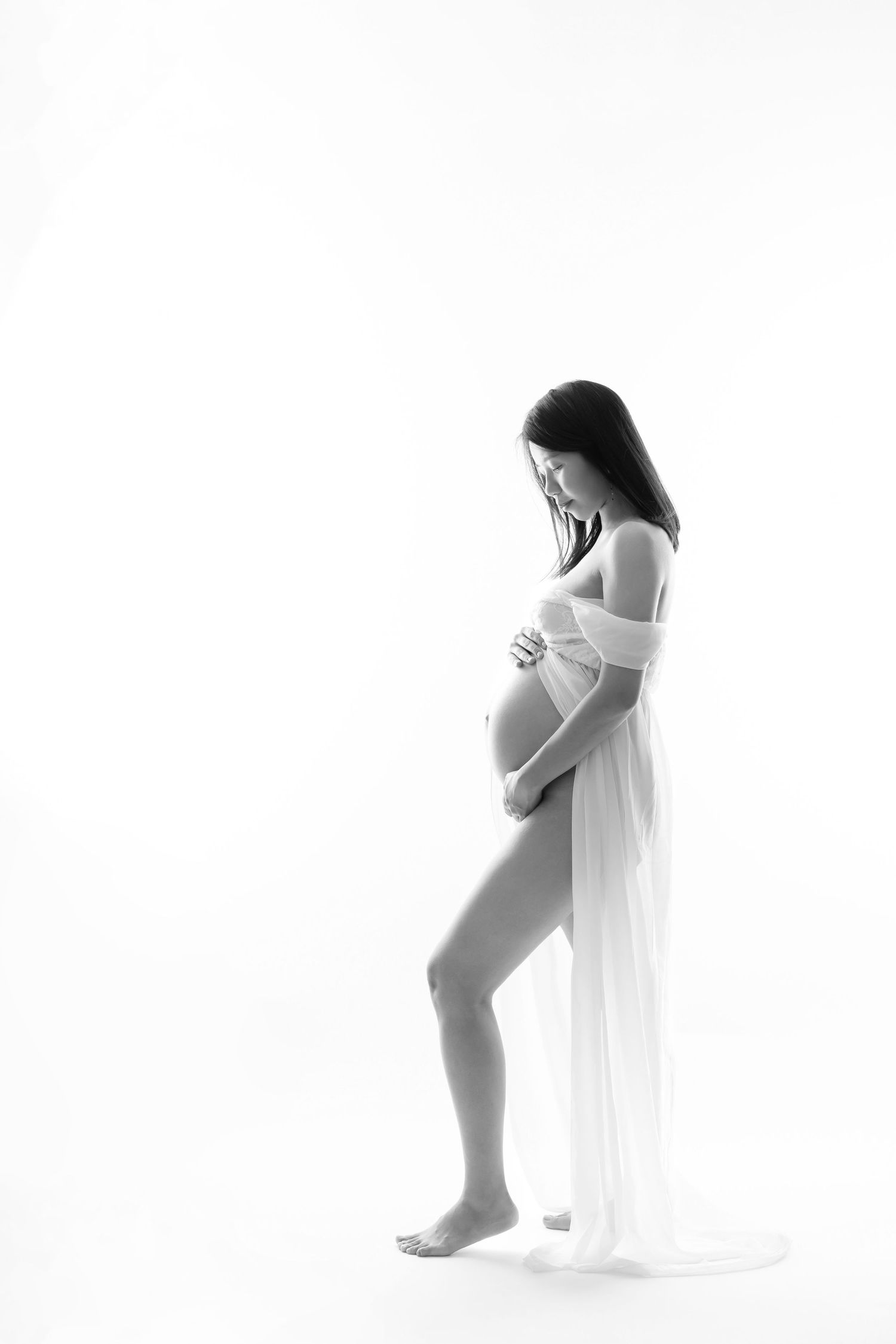 Artistic black and white maternity photo showing an expectant figure draped in flowing white fabric against bright background.