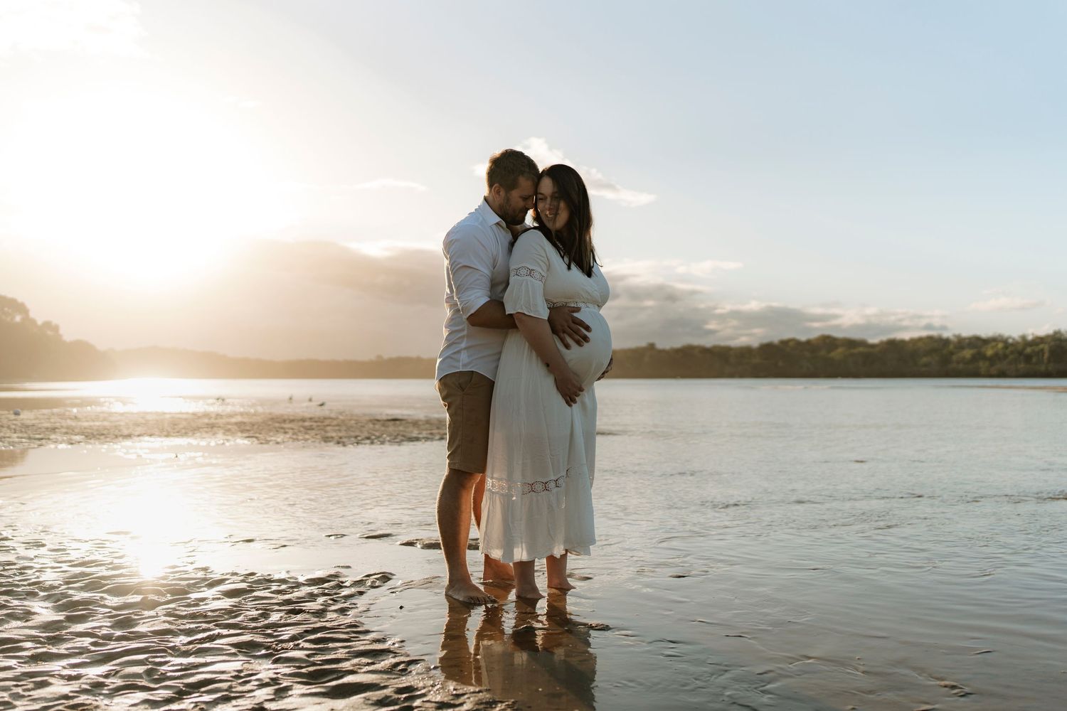 Two people embrace on a beach at sunset with mountains and water in the background creating a romantic silhouette.