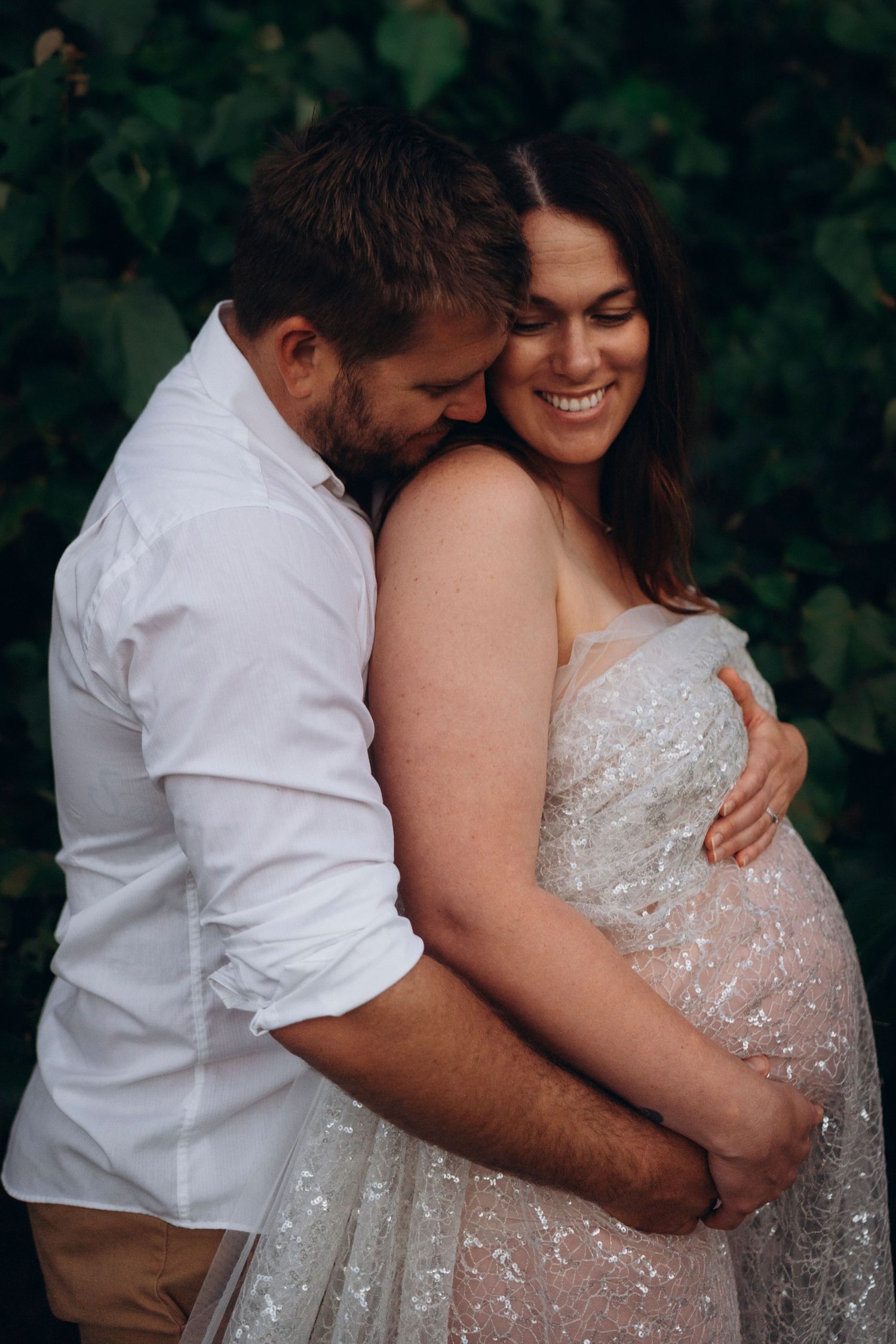 A tender maternity photo outdoors showing couple embracing in white lace dress and casual attire against greenery.