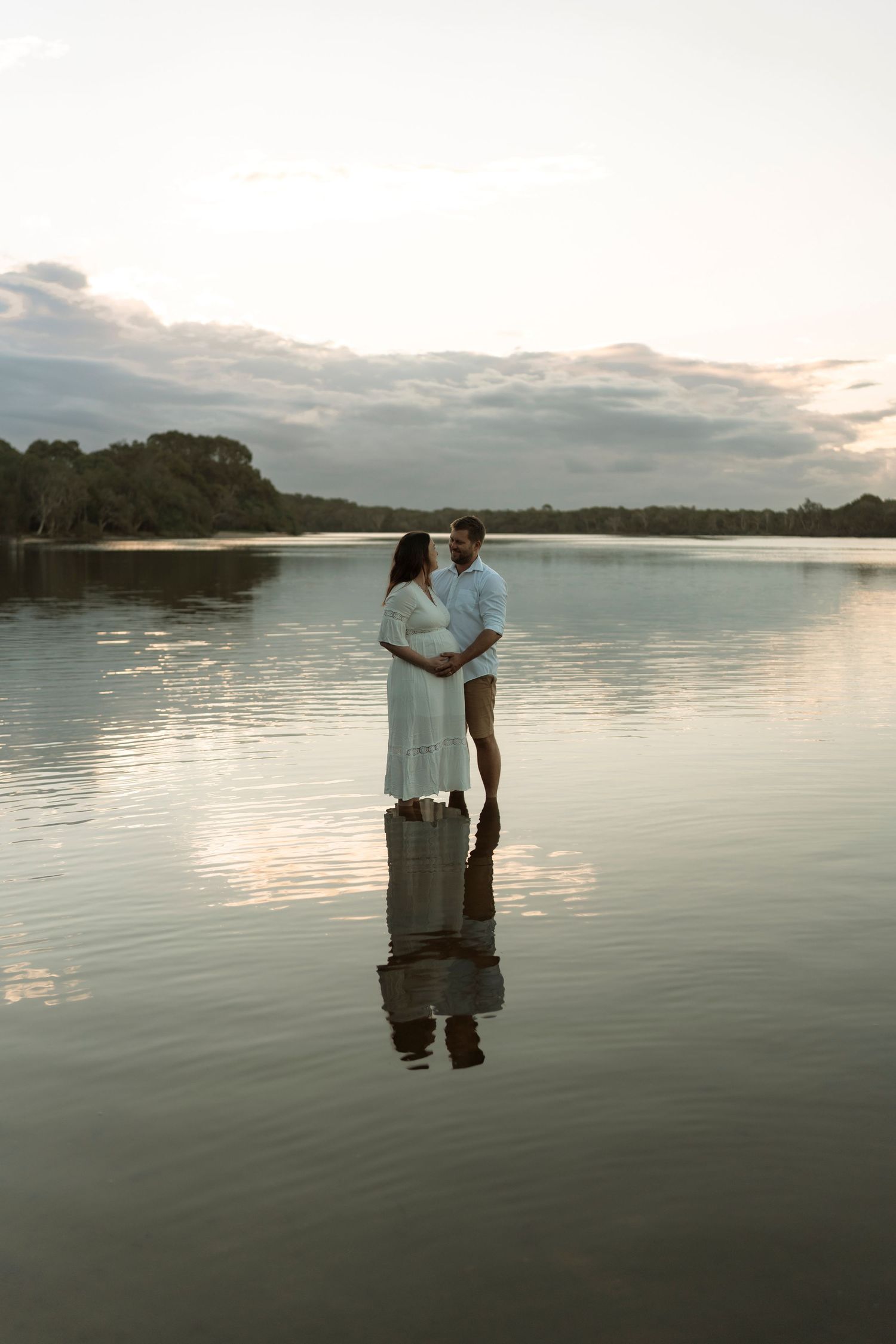 A couple embraces while standing in calm waters during sunset, creating a romantic silhouette reflection on the lake surface.