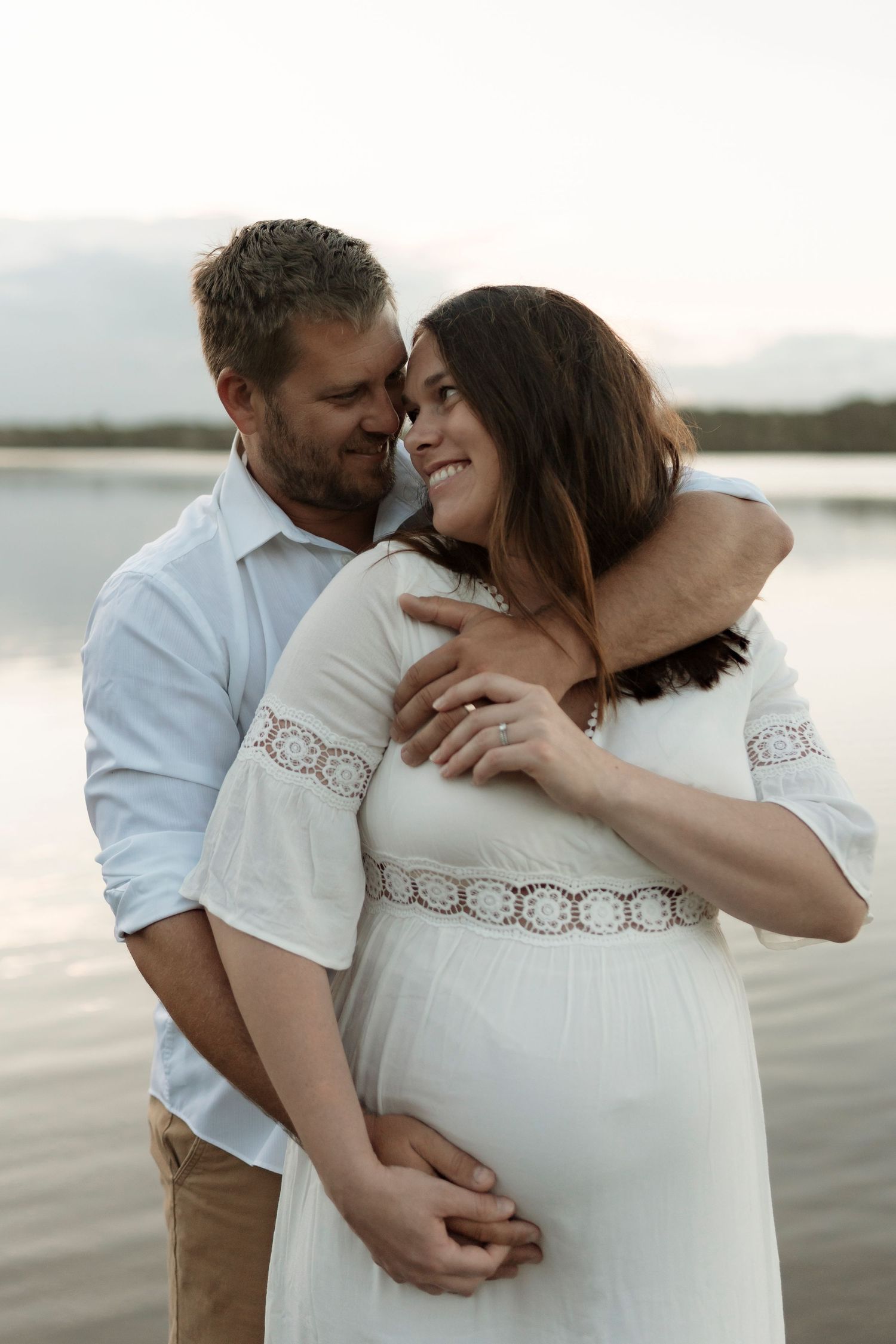 A couple shares an intimate embrace on a beach at sunset, wearing a white dress and casual attire.
