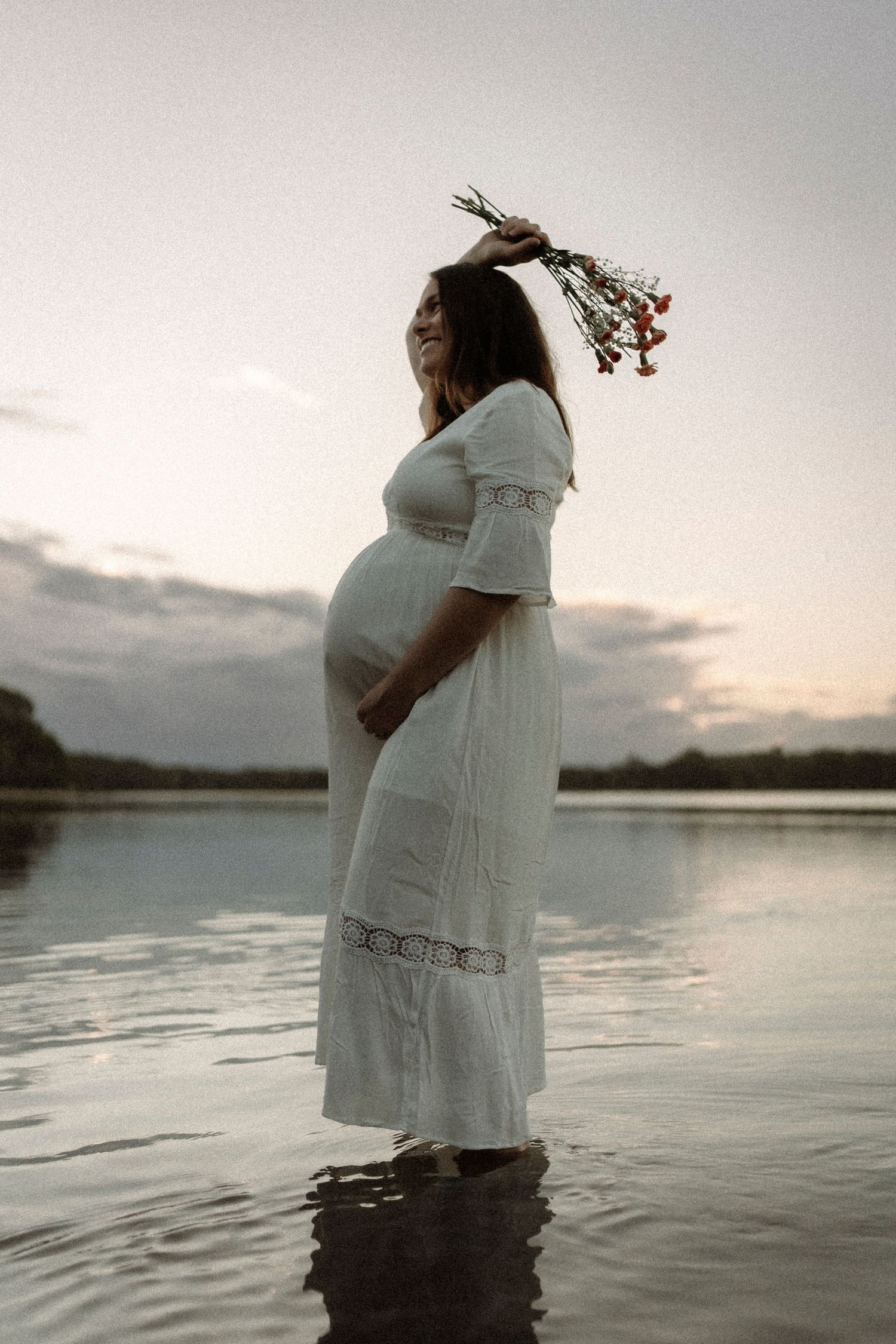 A person in a white maxi dress stands ankle-deep in calm water at sunset holding flowers above their head.