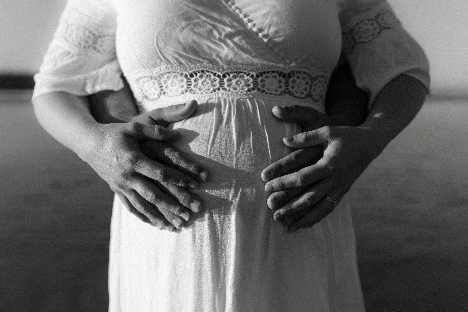 Black and white close-up of hands cradling a pregnant belly in a flowing white maternity dress while standing.