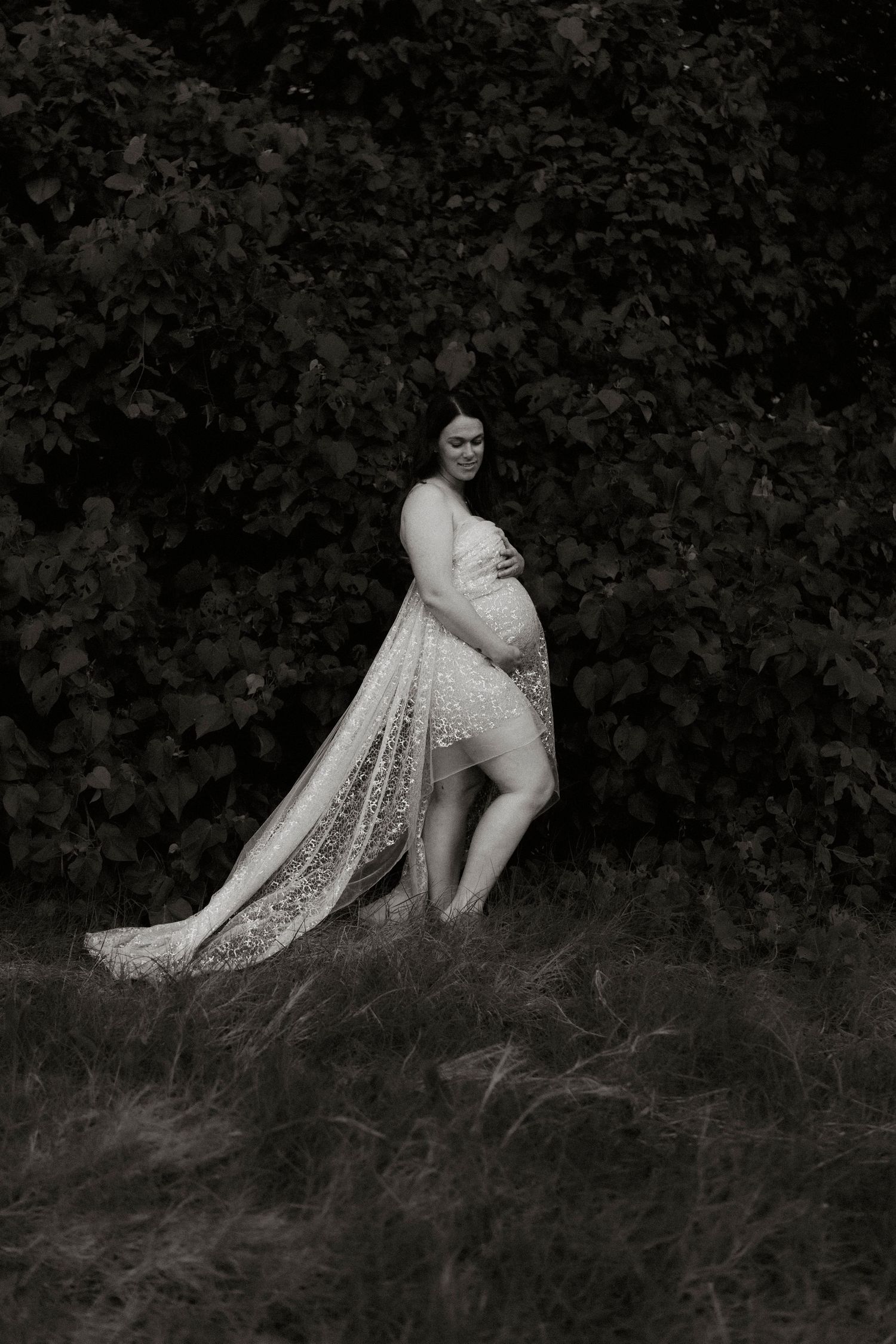 Black and white maternity photography series featuring a pregnant woman in a lace dress reclining on grassy terrain.