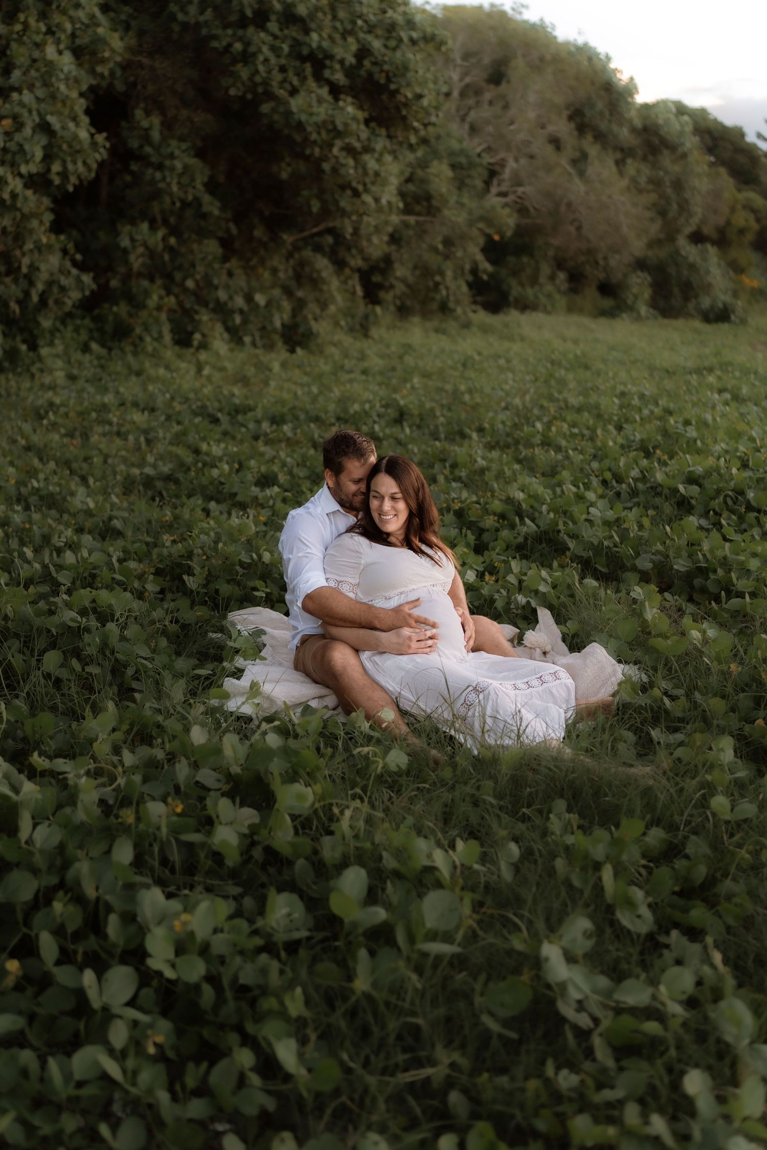 Two people in white outfits share an intimate moment while laying together among lush green vines in a serene outdoor setting.